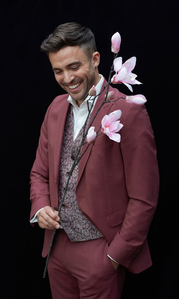 Ein Mann in einem rötlichen Anzug lächelt, während er einen Zweig mit zarten rosa Magnolien in der Hand hält. Der Hintergrund ist schwarz.
