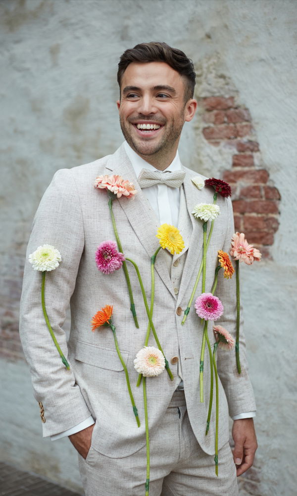 Ein fröhlicher Mann in einem hellen Anzug mit aufgesteckten Gerbera-Blüten in Pink, Gelb und Orange, vor einer Wand mit sichtbaren Ziegelsteinen.