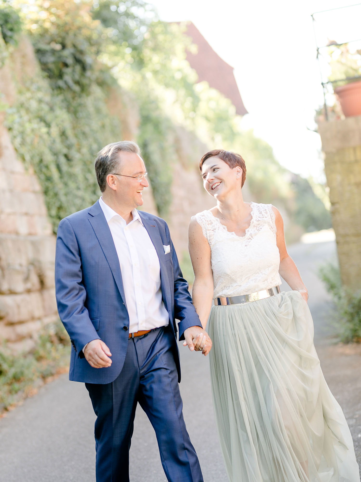Fröhliches Paar in festlicher Kleidung, das zusammen durch eine von Mauern und Bäumen gesäumte Gasse spaziert. Der Mann trägt einen marineblauen Anzug, die Frau einen leichten, pastellfarbenen Rock und ein weißes Oberteil mit Spitze.