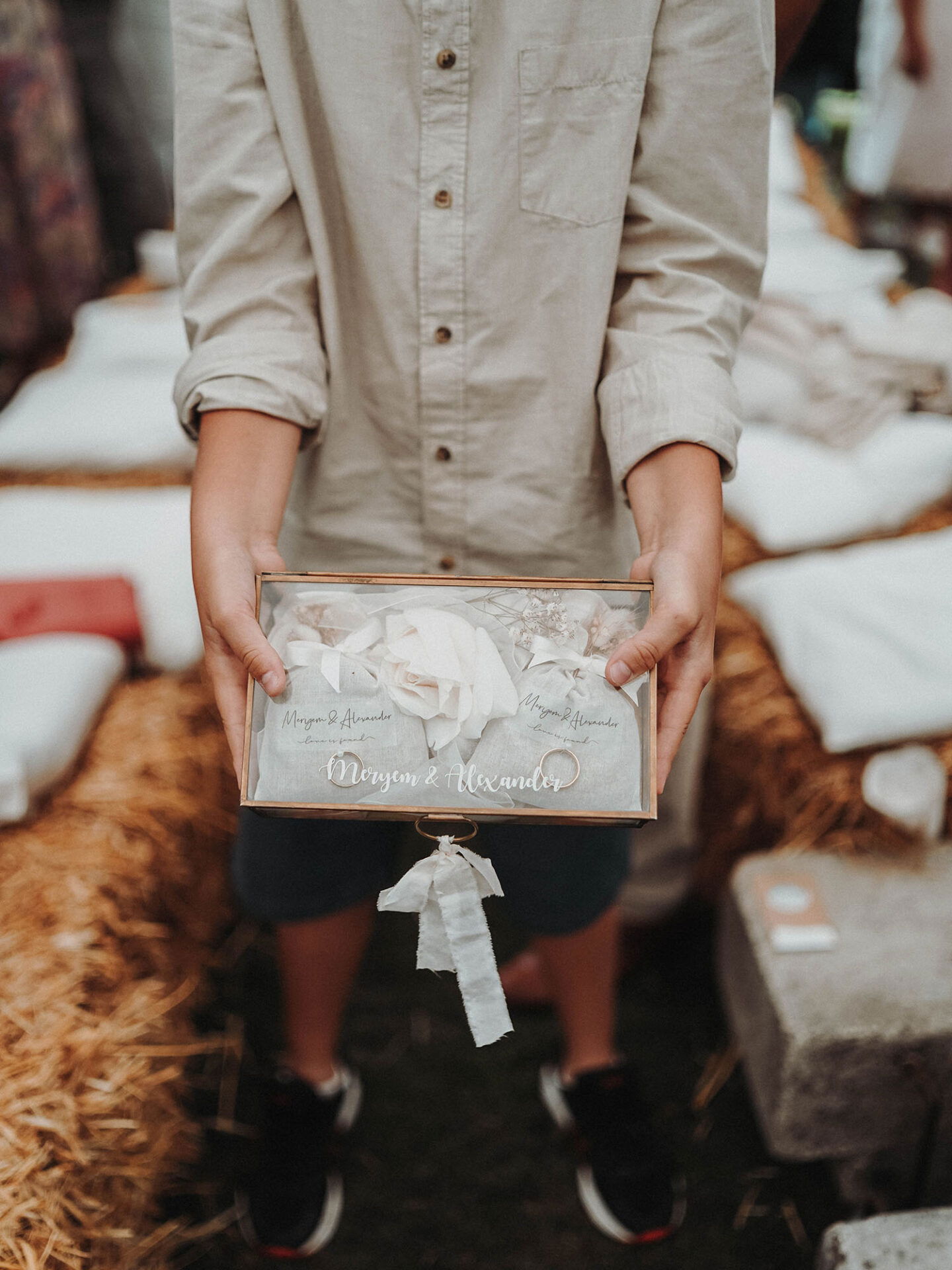 Nahaufnahme von einem Kind, das eine stilvolle Box mit der Aufschrift 'Meryem & Alexander' hält, die mit Schleifen und floralen Akzenten verziert ist. Im Hintergrund sind Strohballen als Teil der Hochzeitsdekoration zu erkennen.