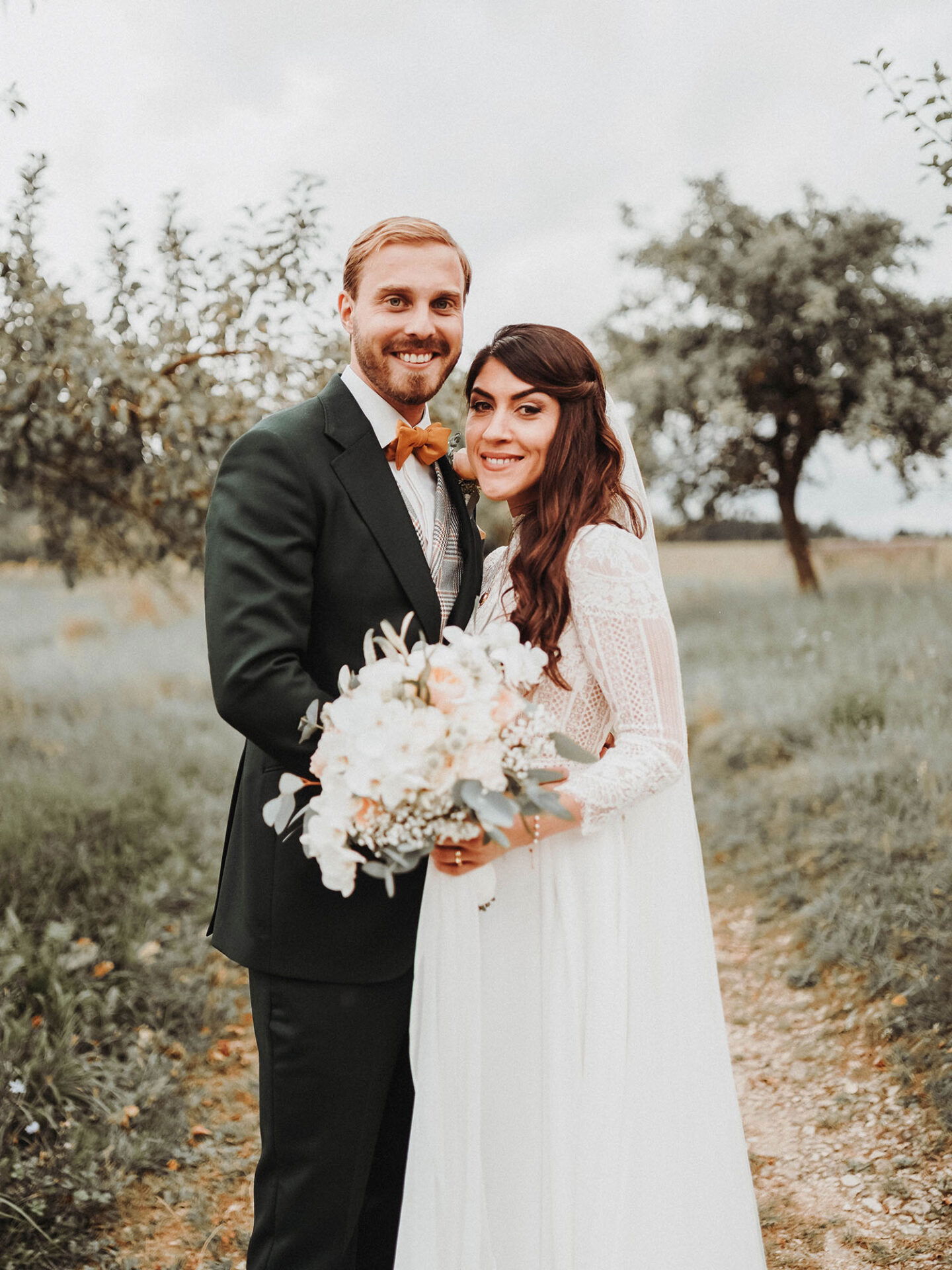 Das Hochzeitspaar, Meryem und Alexander, posiert inmitten einer natürlichen Landschaft. Der Bräutigam trägt einen eleganten grünen Anzug mit einem gelben Akzent, während die Braut ein romantisches Spitzenkleid und einen Blumenstrauß trägt.