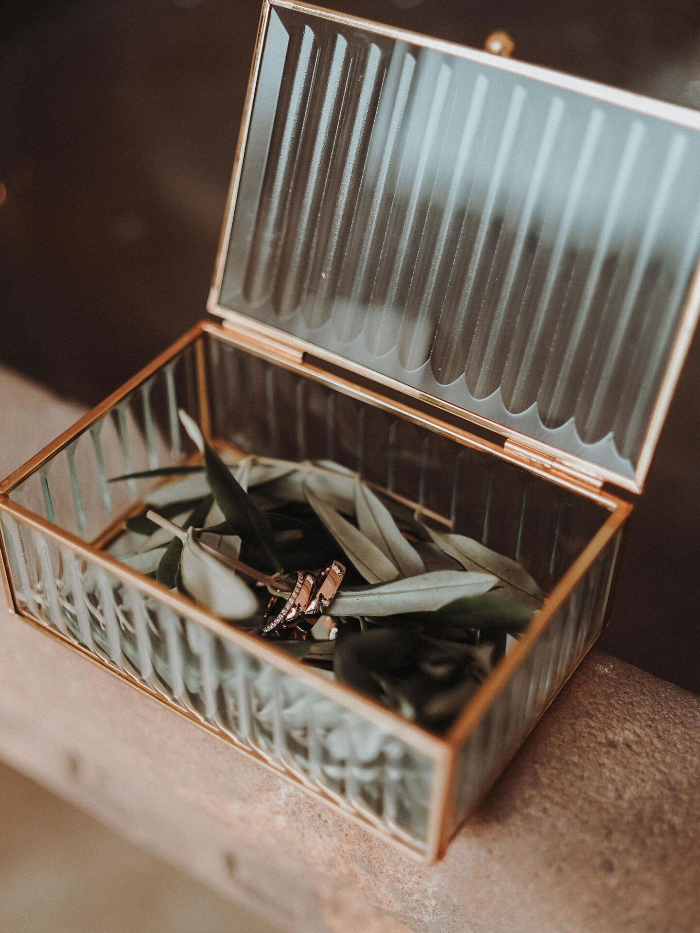 Eine filigrane Glasbox mit goldenem Rahmen, in der auf Olivenblättern Eheringe liegen, ist geöffnet und auf einer Steinoberfläche platziert. Die schlichten Ringe kontrastieren harmonisch mit der natürlichen Dekoration.