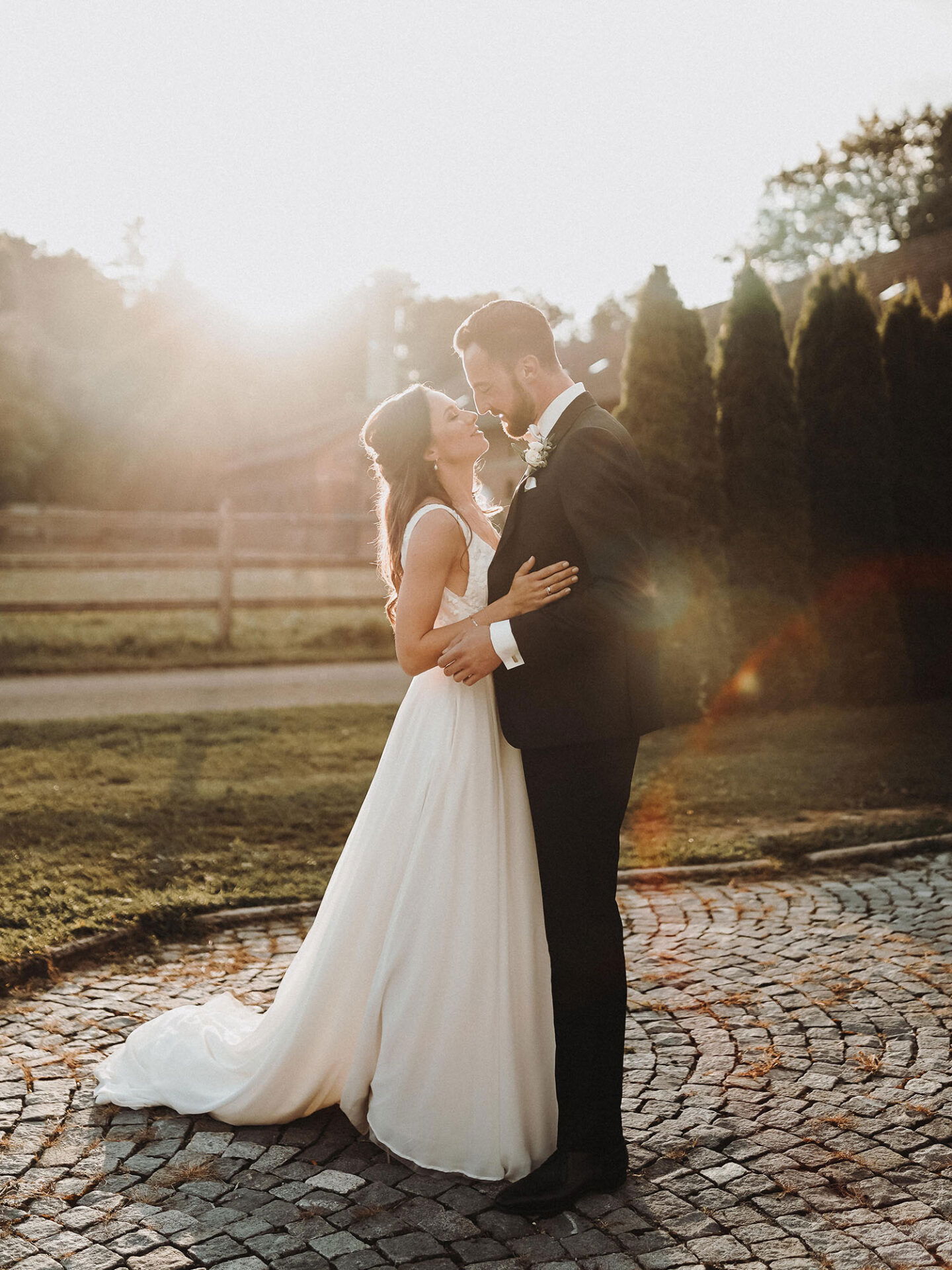 Ein Hochzeitspaar genießt einen innigen Moment im Freien. Die Braut, in einem langen weißen Kleid, lächelt ihren Bräutigam an, der in einem klassischen dunklen Anzug gekleidet ist. Die Szene wird von der untergehenden Sonne stimmungsvoll erhellt.