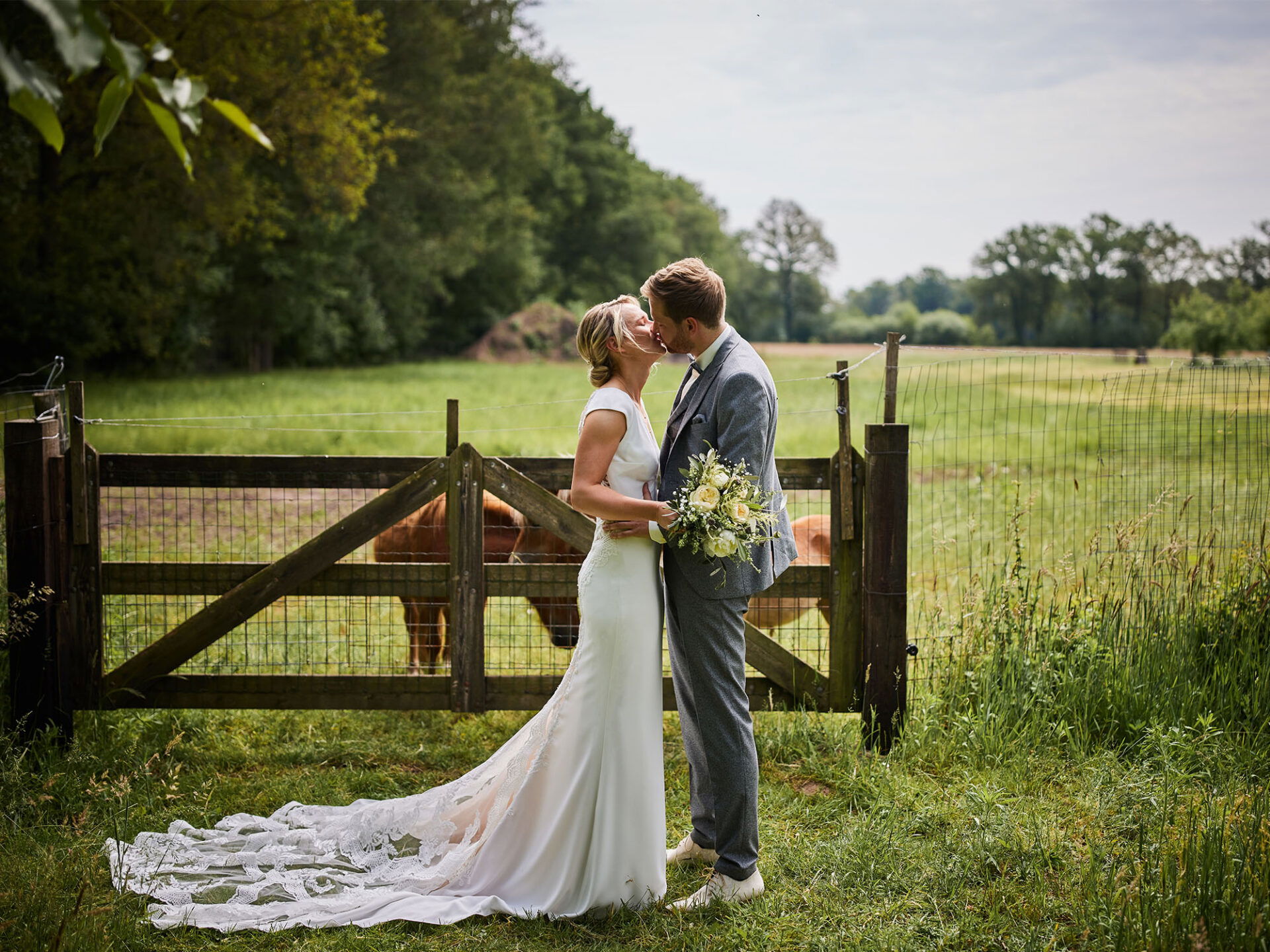 Ein Hochzeitspaar steht vor einem rustikalen Holztor und küsst sich auf einer Wiese. Die Braut trägt ein langes Kleid mit Schleppe, während der Bräutigam in einem grauen Anzug gekleidet ist. Im Hintergrund befinden sich weidende Kühe.