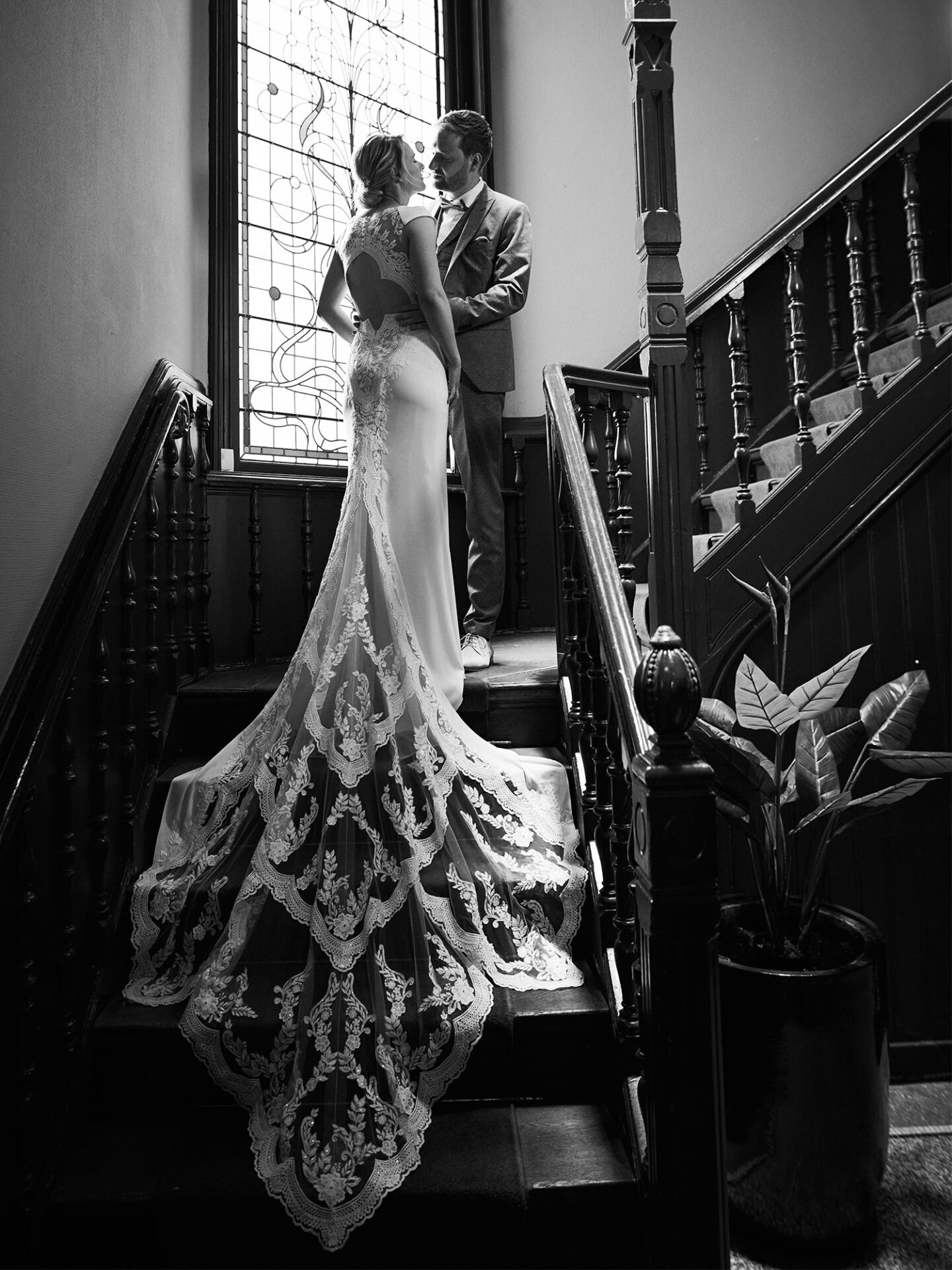 Ein Brautpaar posiert auf einer alten Holztreppe, die Braut in einem Kleid mit langer, kunstvoller Schleppe aus Spitze. Das Paar steht vor einem großen Fenster mit Buntglasdetails. Das Bild ist in Schwarz-Weiß aufgenommen und strahlt eine romantische Stimmung aus.