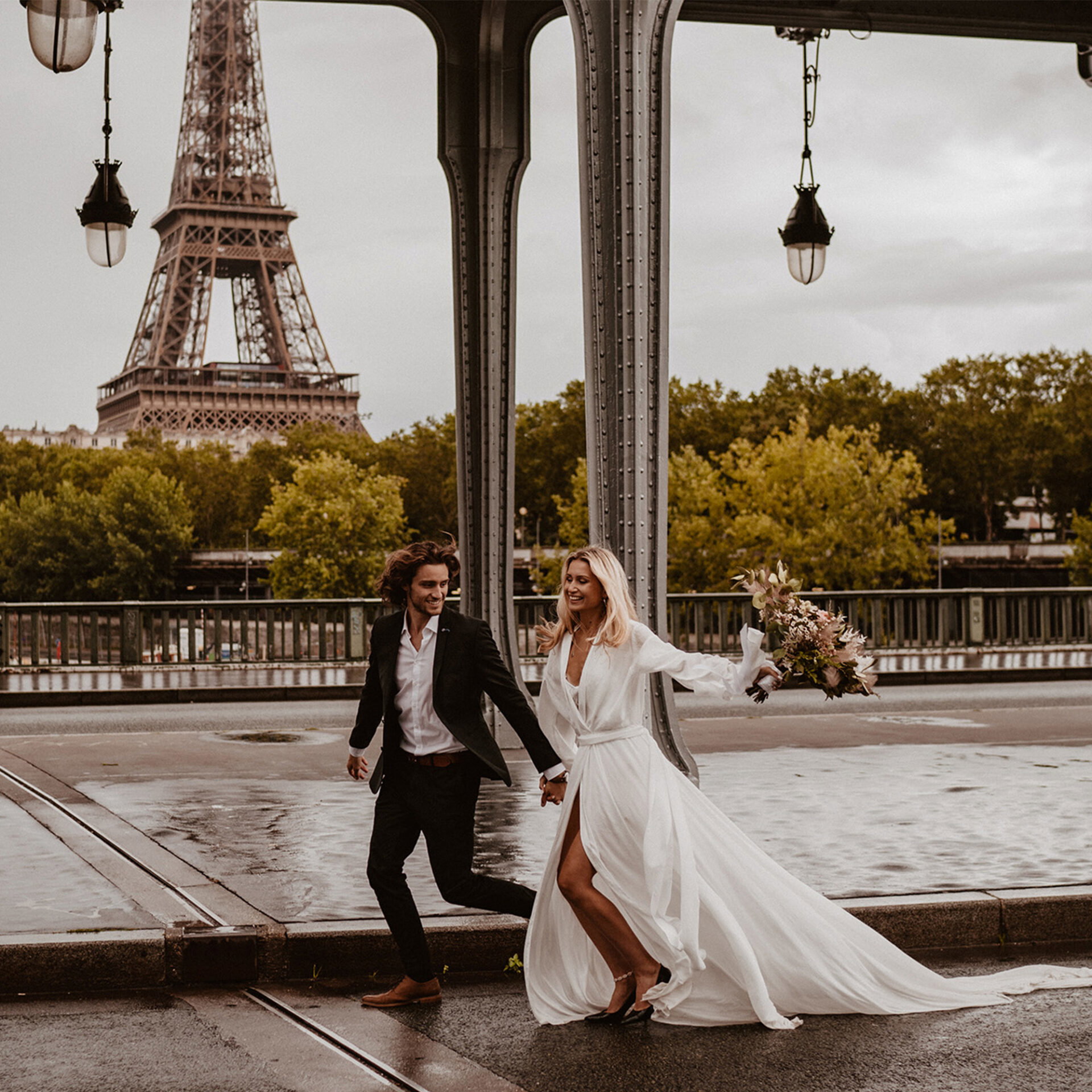 Ein Paar, sie in einem weißen Hochzeitskleid und er im Anzug, läuft fröhlich Hand in Hand auf einer Brücke in Paris mit dem Eiffelturm im Hintergrund.