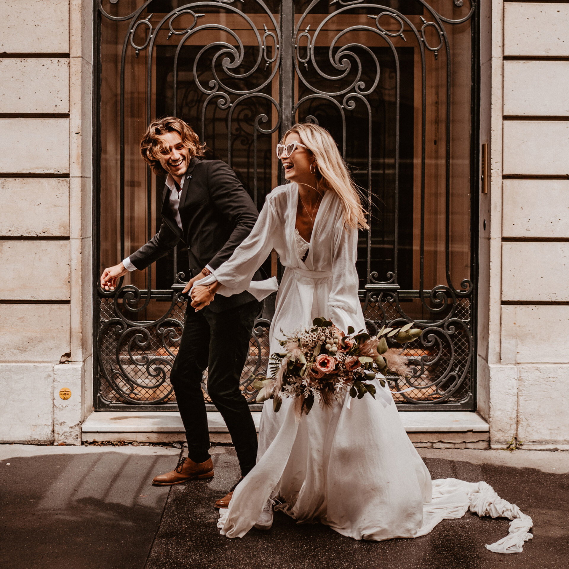 Das Paar, die Braut in einem weißen Kleid und mit Blumenstrauß, der Bräutigam im Anzug, läuft gemeinsam und lachend an einem historischen Gebäude in Paris vorbei.