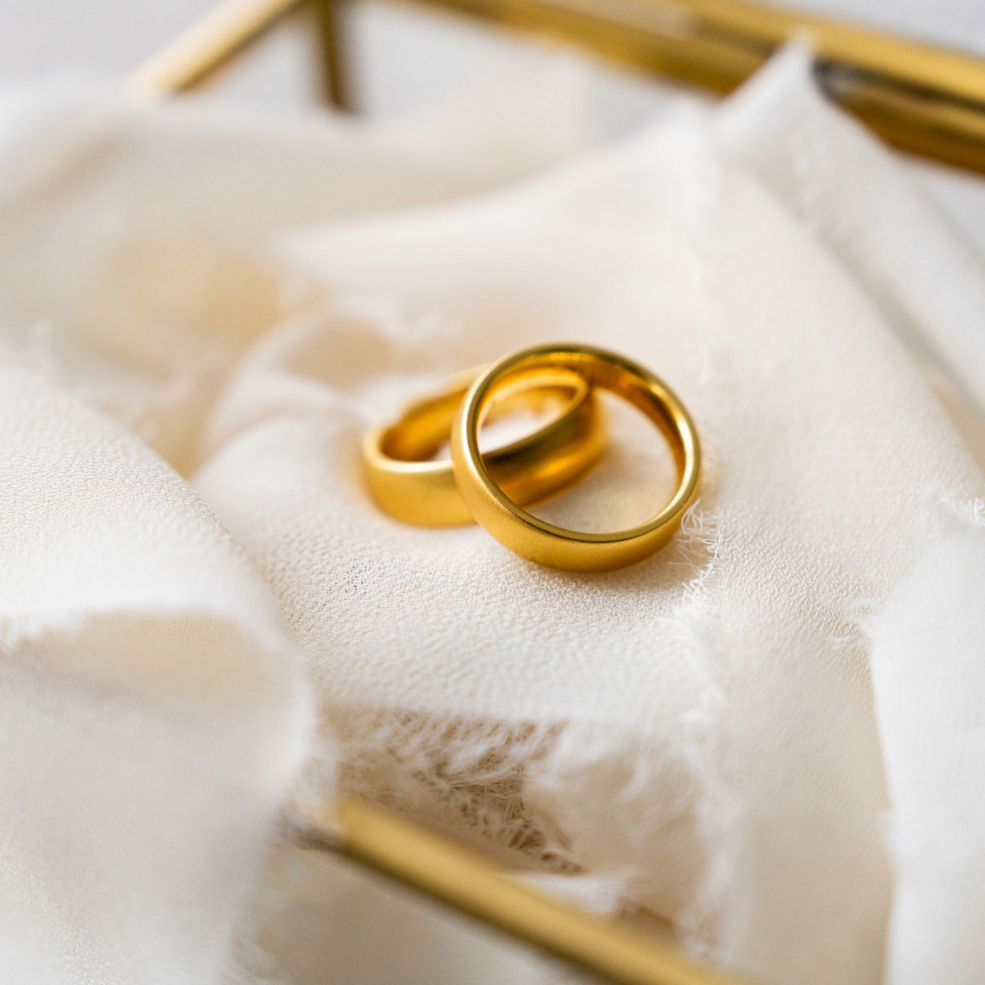 Zwei goldene Trauringe liegen auf einem fransigen, cremefarbenen Stoff in einer stilvollen Glasschatulle. Ein minimalistisches, aber edles Design für die Ringpräsentation.