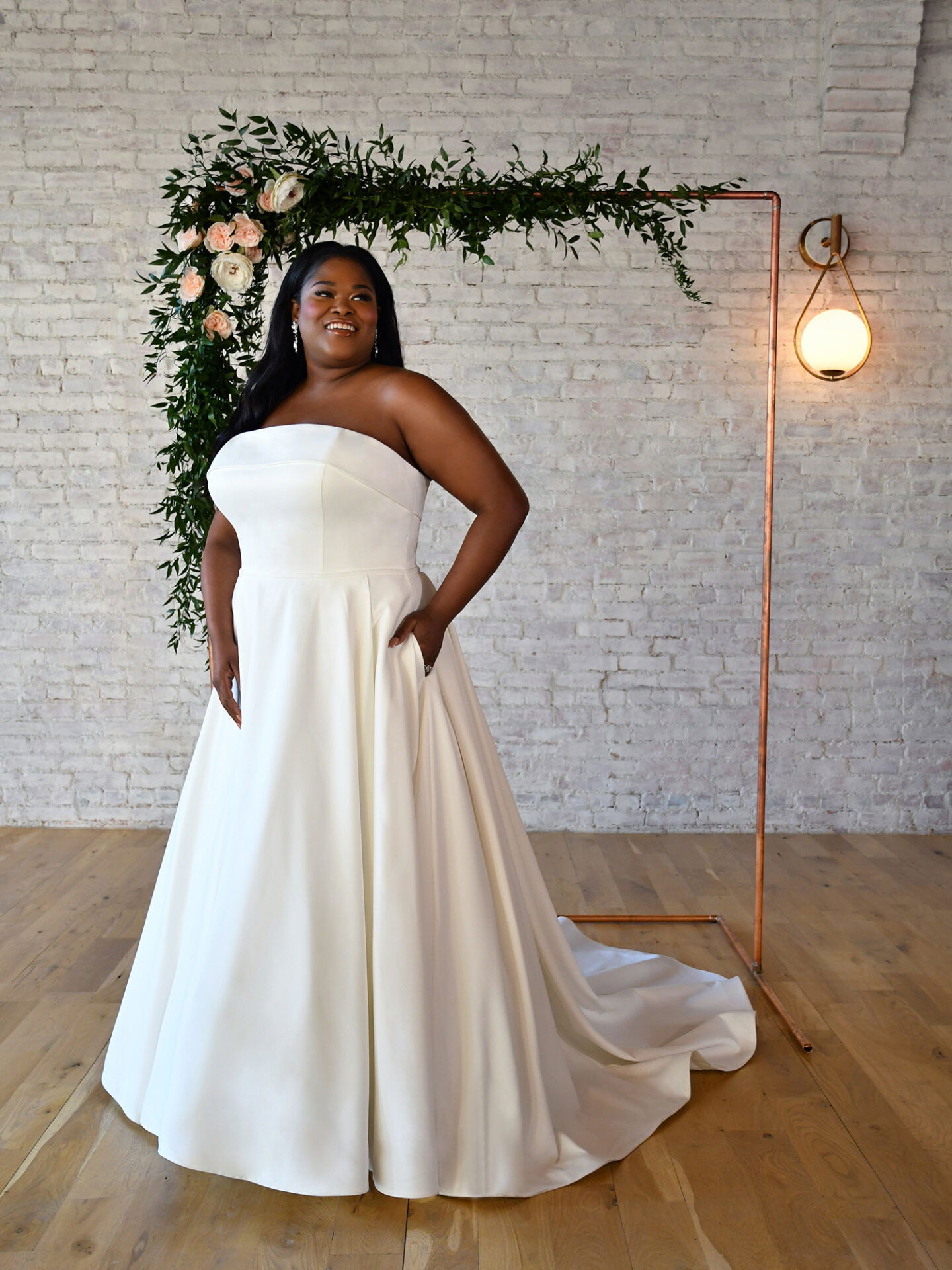 Eine lächelnde Braut in einem schlichten, ärmellosen A-Linien-Hochzeitskleid steht auf einem Holzfußboden, flankiert von einem Kupferrahmen, der mit grünen Zweigen und Rosen dekoriert ist.