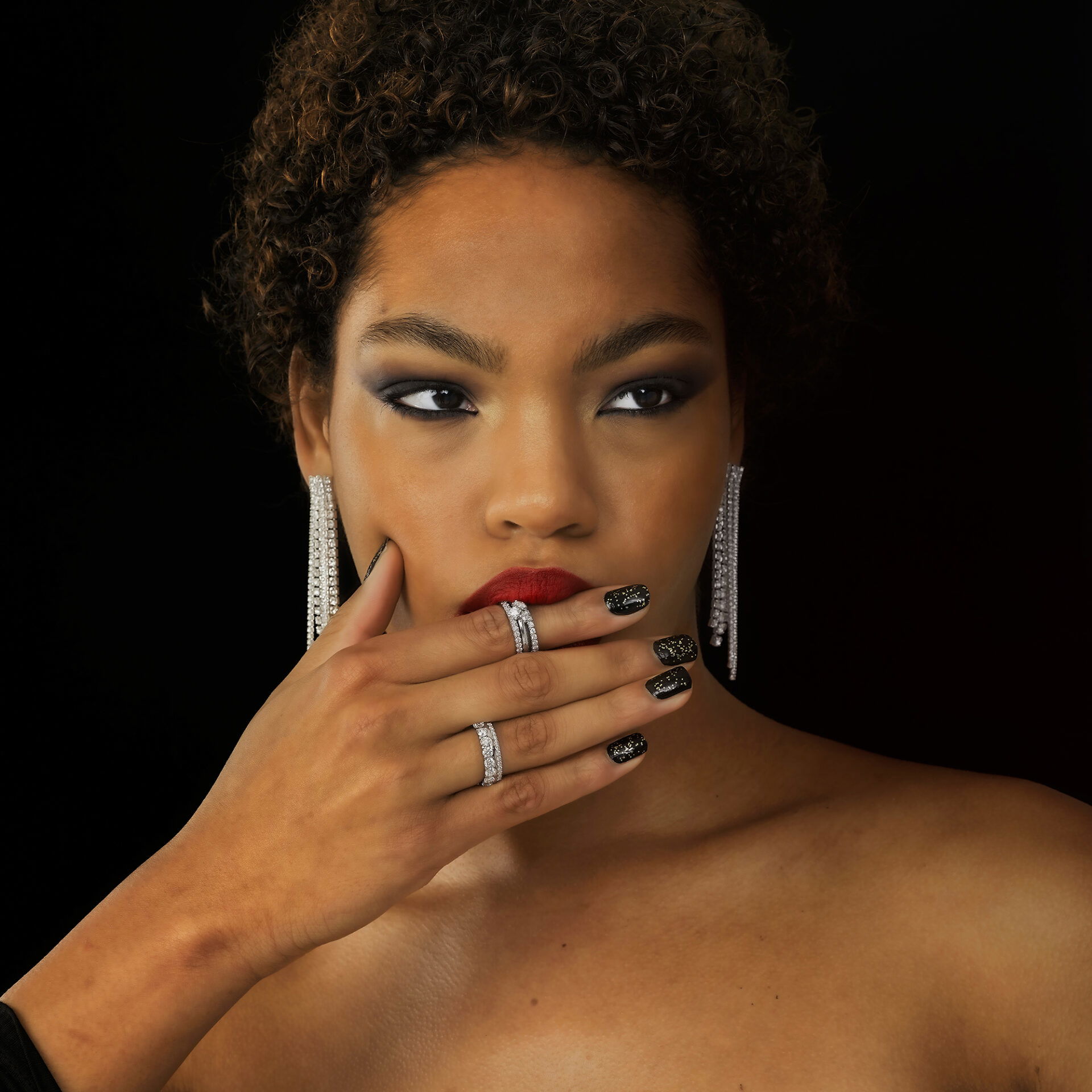 Nahaufnahme einer Frau mit kurzen, lockigen Haaren, die stilvolle, lange Ohrringe und funkelnde Ringe trägt. Eine Hand mit schwarzem Nagellack liegt leicht auf ihrem Gesicht und sorgt für eine mystische Atmosphäre.