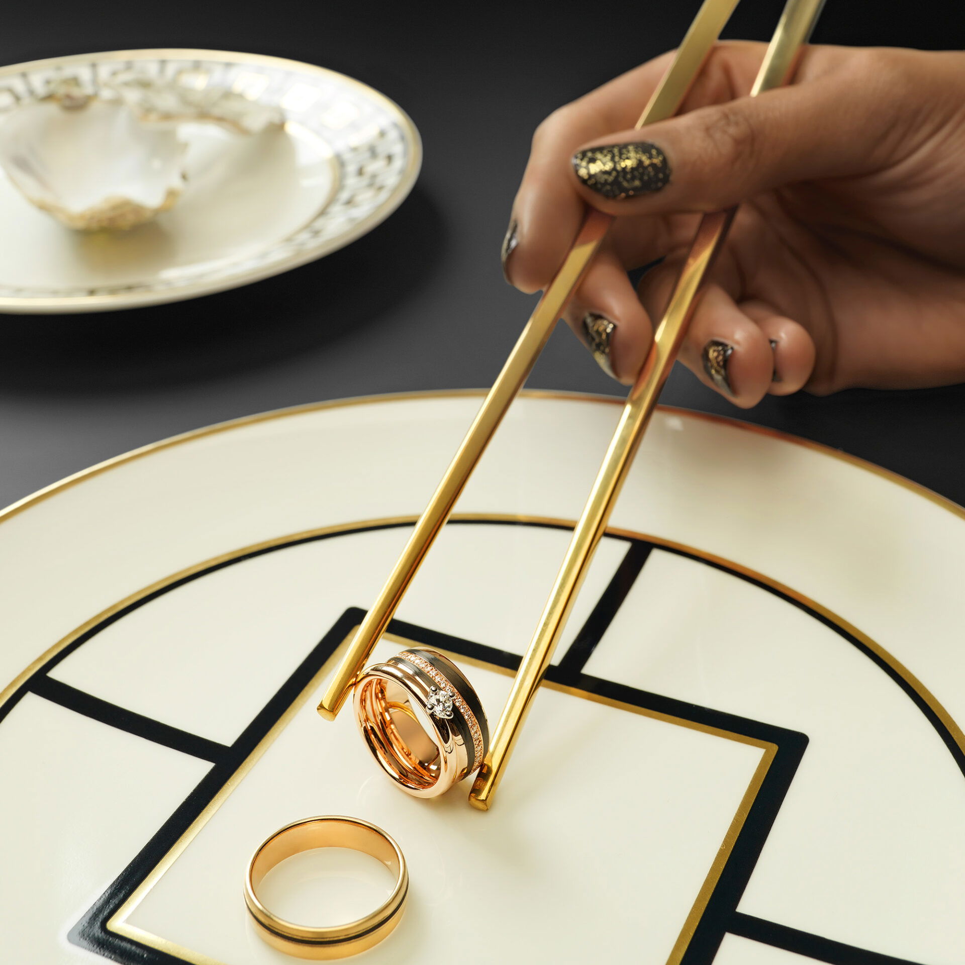 Ein goldener Ehering wird mit Essstäbchen angehoben, während ein weiterer Ring auf einem kunstvoll gemusterten Teller liegt. Ein modernes und minimalistisches Design.