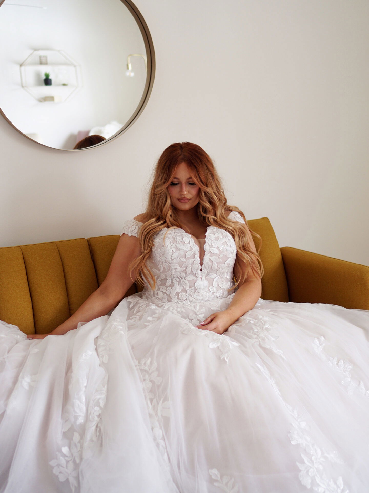 Brautmodell in einem voluminösen, mit Spitze verzierten Brautkleid sitzt auf einem senfgelben Sofa in einem stilvollen, minimalistisch eingerichteten Raum mit einem runden Spiegel im Hintergrund.