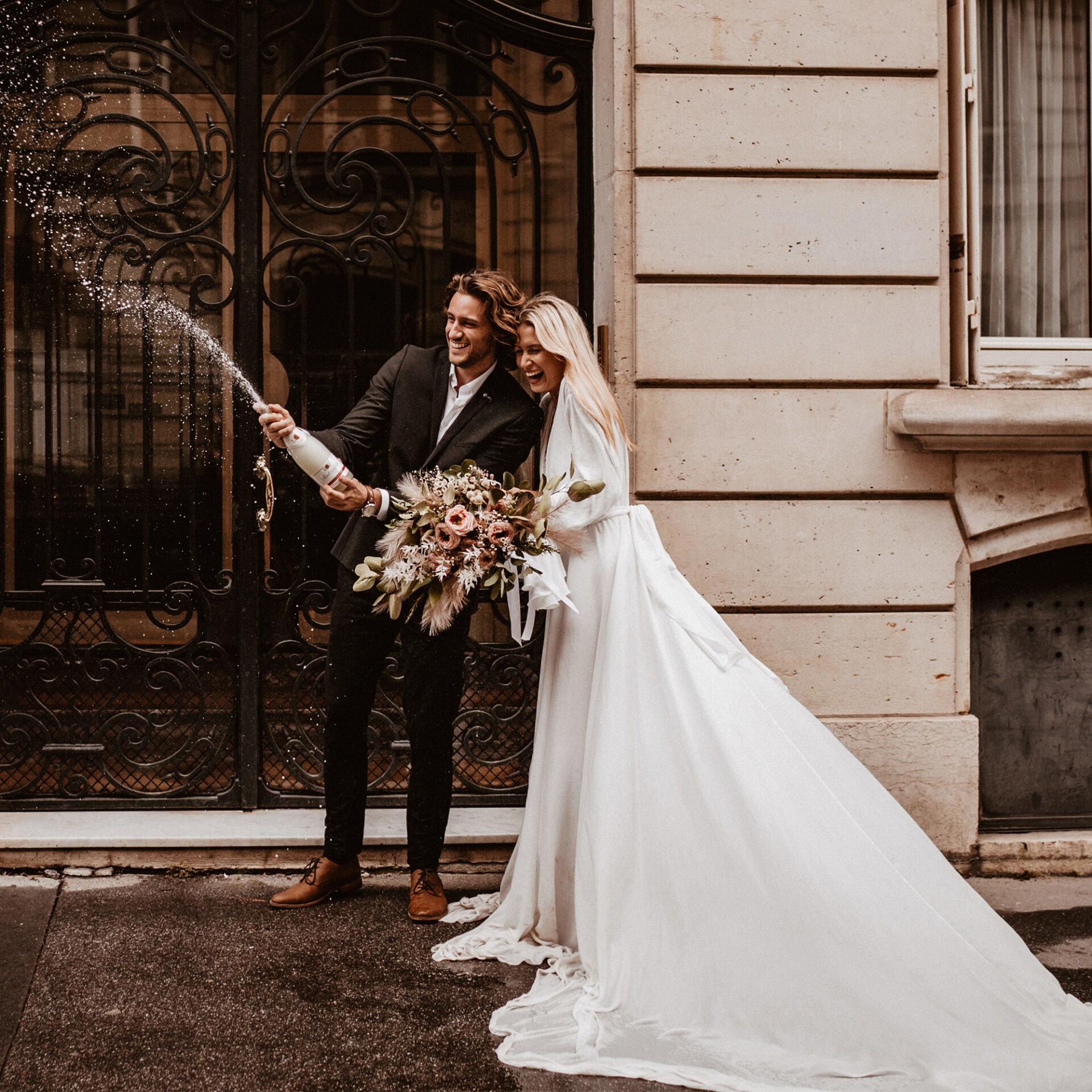 Ein lachendes Brautpaar steht vor einer kunstvoll verzierten Tür in Paris. Der Bräutigam spritzt Champagner, während die Braut einen großen Blumenstrauß hält.