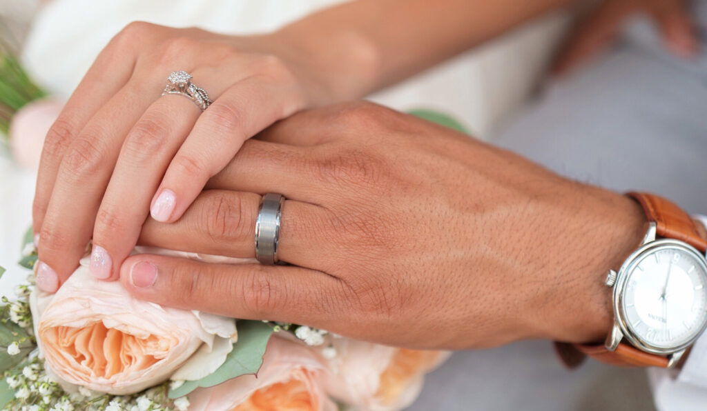 Hände eines frisch verheirateten Paares mit Eheringen, die liebevoll übereinander gelegt sind und auf einem rosafarbenen Brautstrauß ruhen, während der Bräutigam eine Armbanduhr trägt.