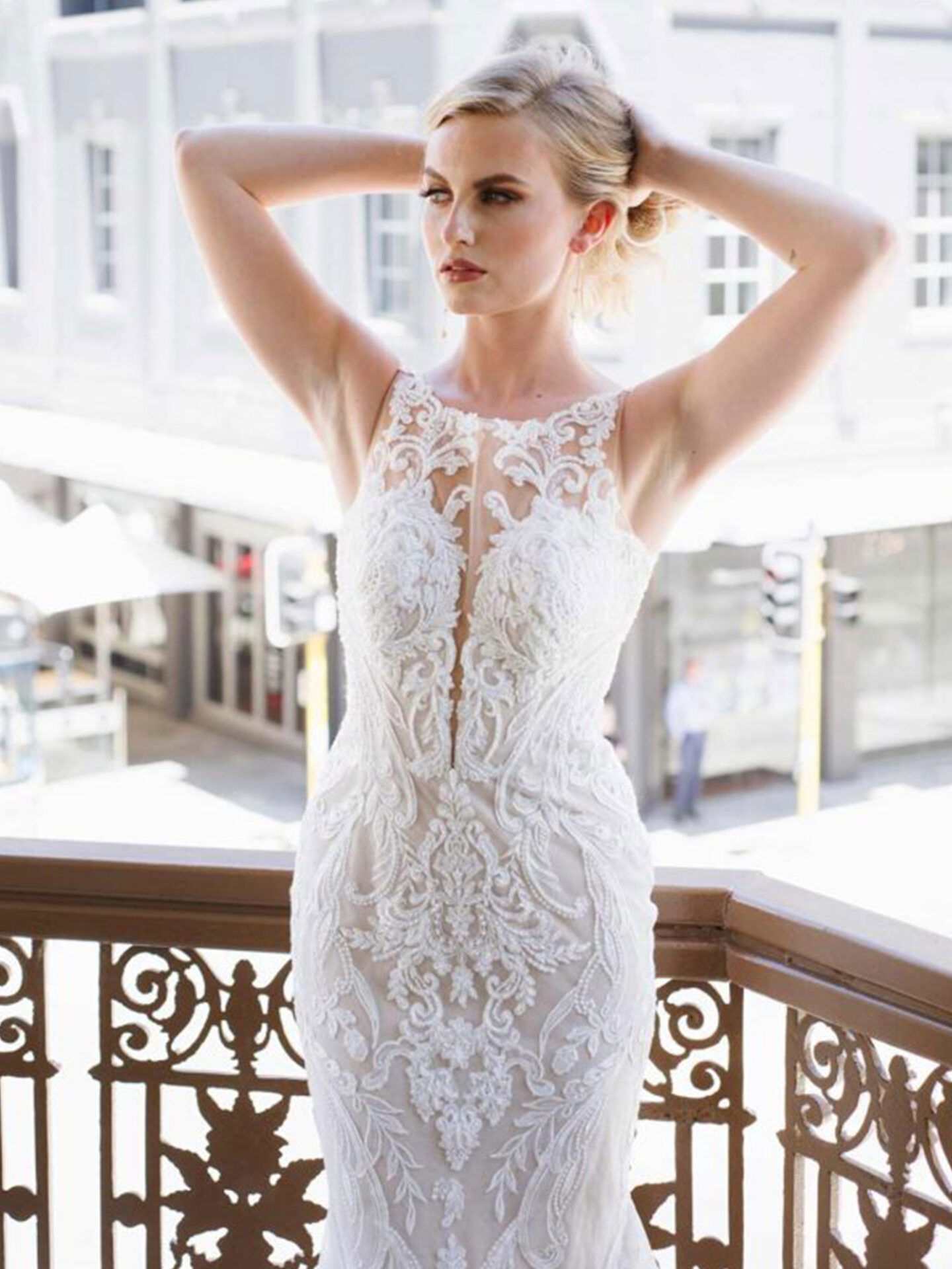 Vordere Ansicht einer Braut in einem ärmellosen Hochzeitskleid mit detaillierten Stickereien, die auf einem Balkon steht, ihre Arme nach oben streckt und durch die Straßen der Stadt schaut.