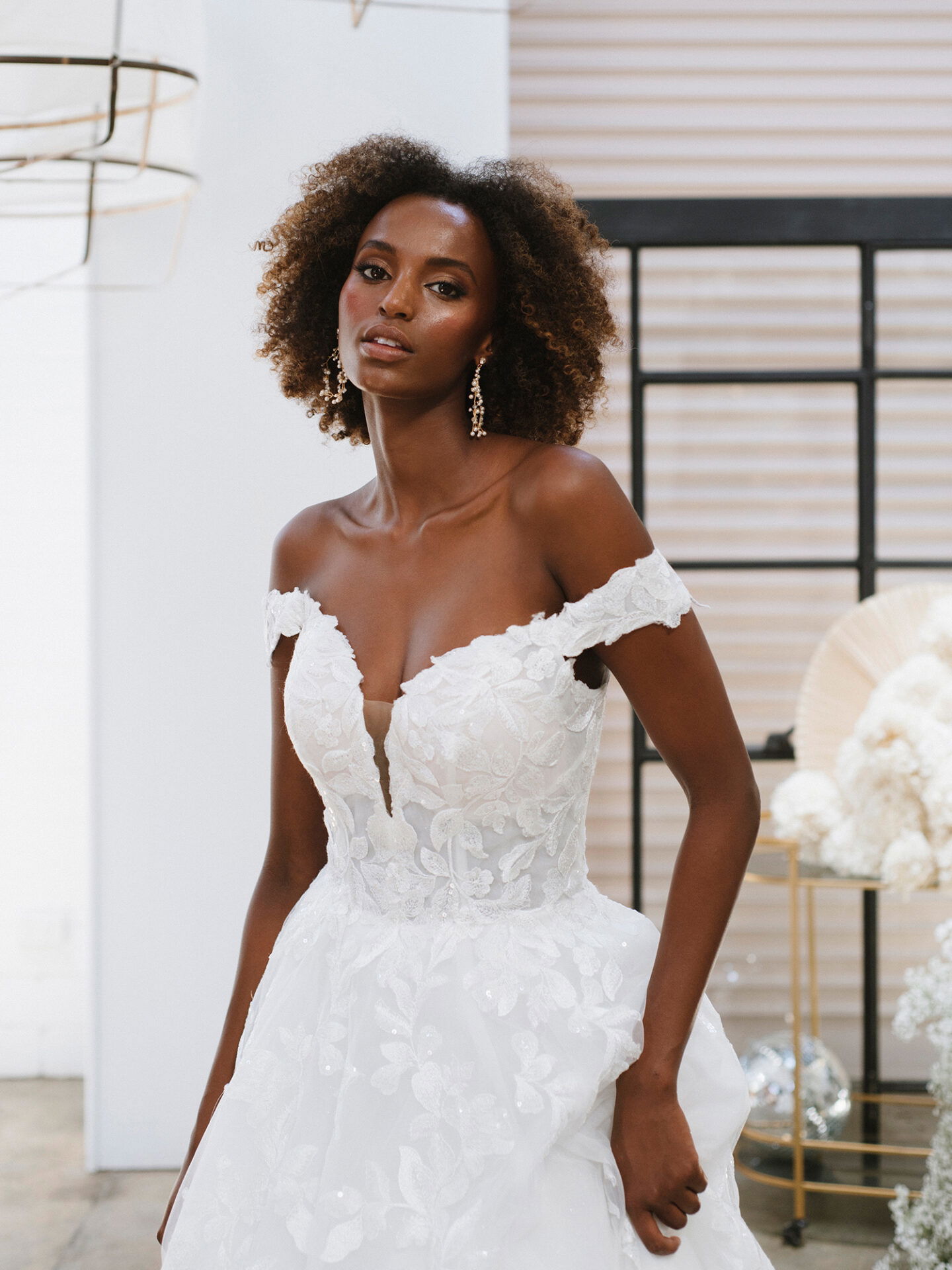 Braut in einem schulterfreien Brautkleid mit romantischen Spitzenverzierungen und tiefem V-Ausschnitt, trägt Perlenohrringe und posiert in einem modernen, lichtdurchfluteten Raum.
