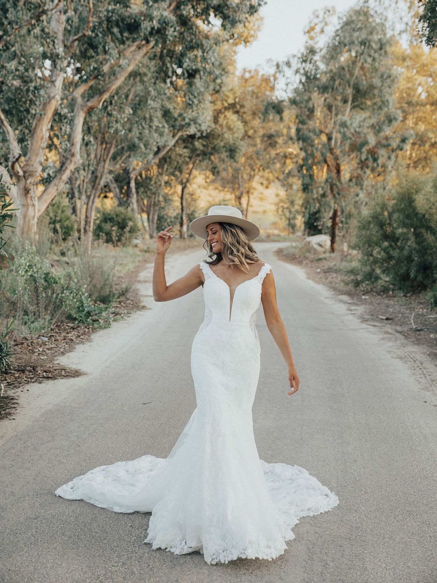 Braut in einem weißen Spitzenkleid mit tiefem Ausschnitt und Hut, die selbstbewusst auf einer verlassenen Straße geht, umgeben von hohen Bäumen und goldenen Sonnenstrahlen.