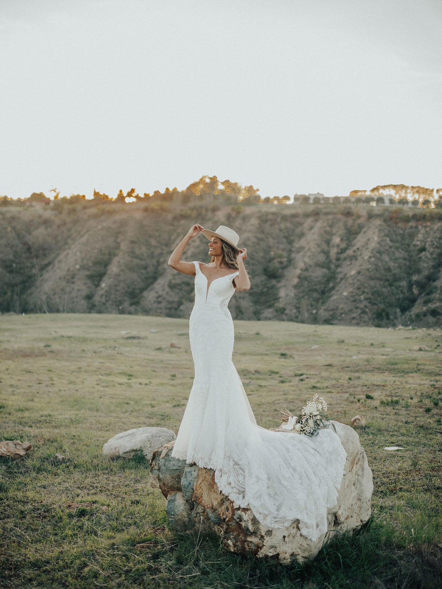 Eine Braut in einem weißen Spitzen-Hochzeitskleid posiert auf einem großen Felsen in einer grasbewachsenen Landschaft, während sie den Hut anhebt und sich im sanften Licht des Sonnenuntergangs sonnt.