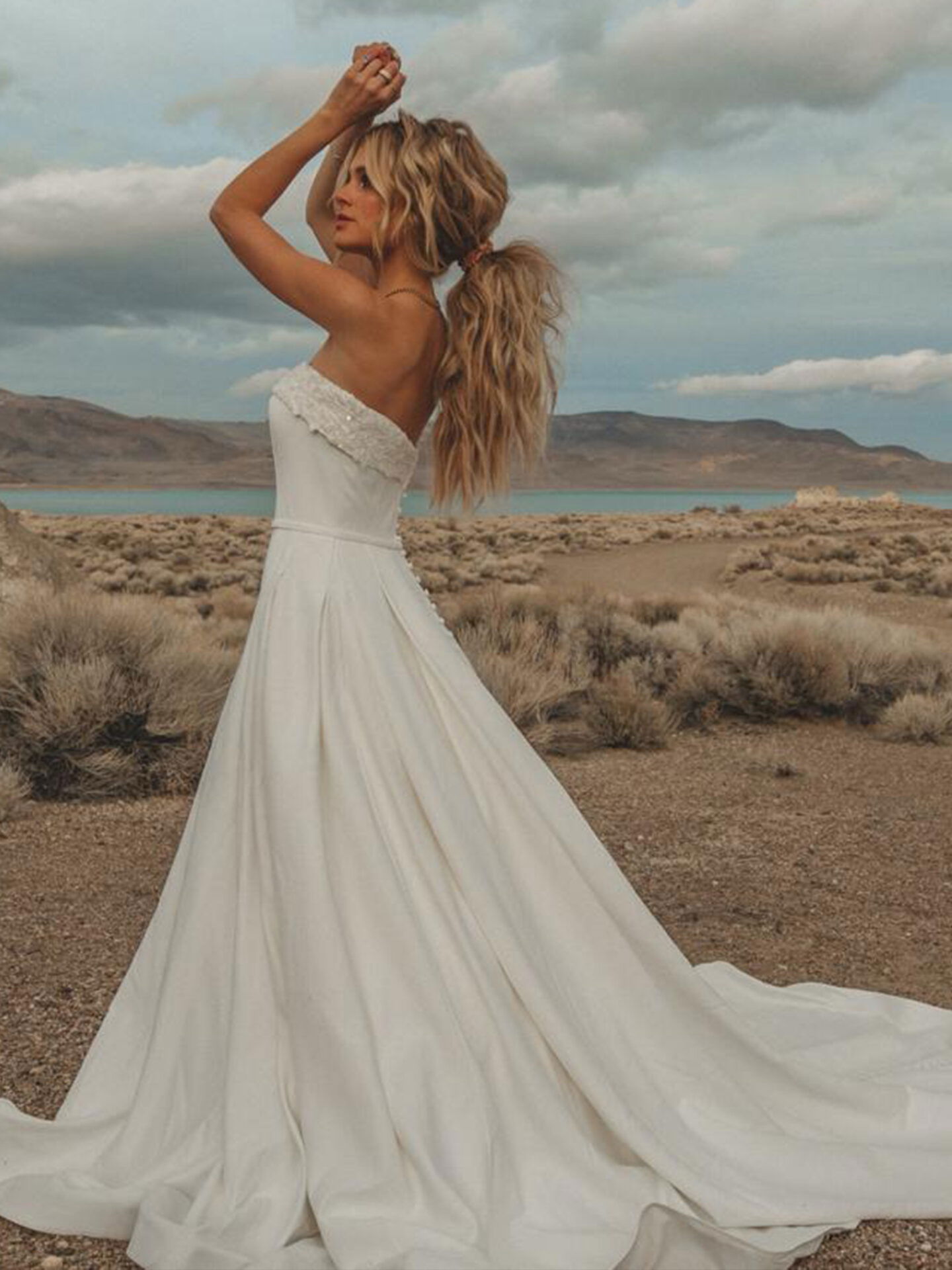 Eine Braut in einem trägerlosen Hochzeitskleid mit einer eleganten Schleppe posiert in einer weiten Wüstenlandschaft und streckt die Arme, während ihr langes, lockiges Haar in den Wind fällt.