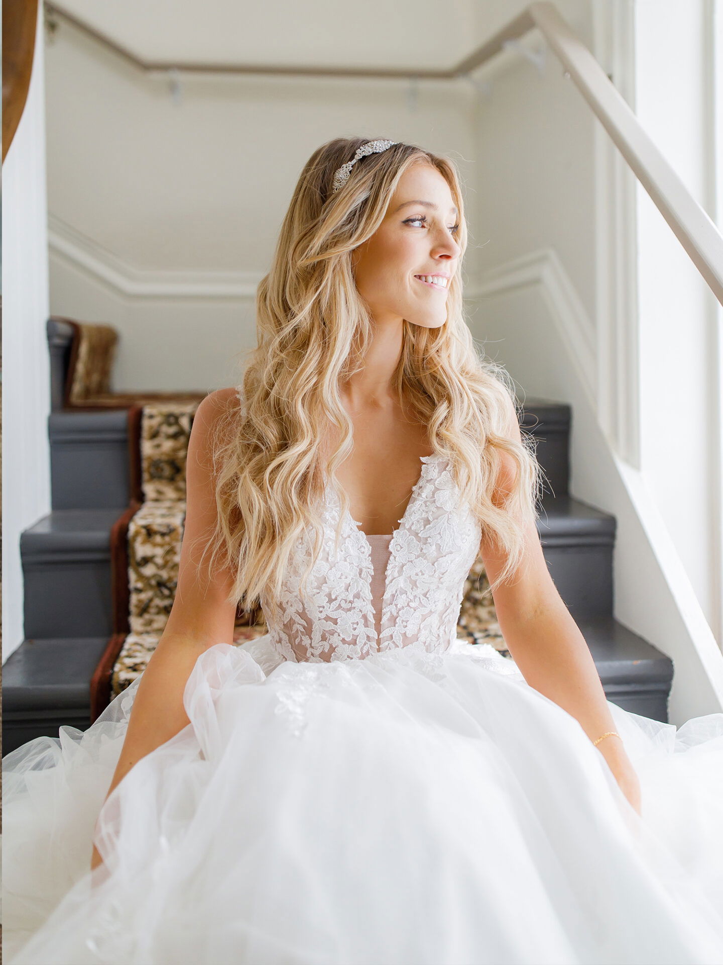 Braut trägt ein Spitzenkleid mit tiefem Ausschnitt und sitzt in einem lichtdurchfluteten Raum auf einer Treppe, ihr welliges Haar und die Spitzenapplikationen ihres Kleides strahlen Eleganz aus.