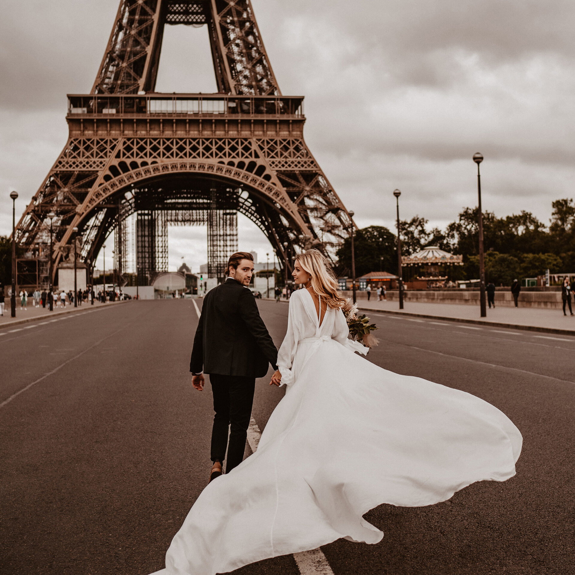 Ein Paar in Hochzeitskleidung läuft auf der Straße vor dem Eiffelturm. Das weiße Kleid der Braut weht im Wind, während der Bräutigam sie lächelnd ansieht.