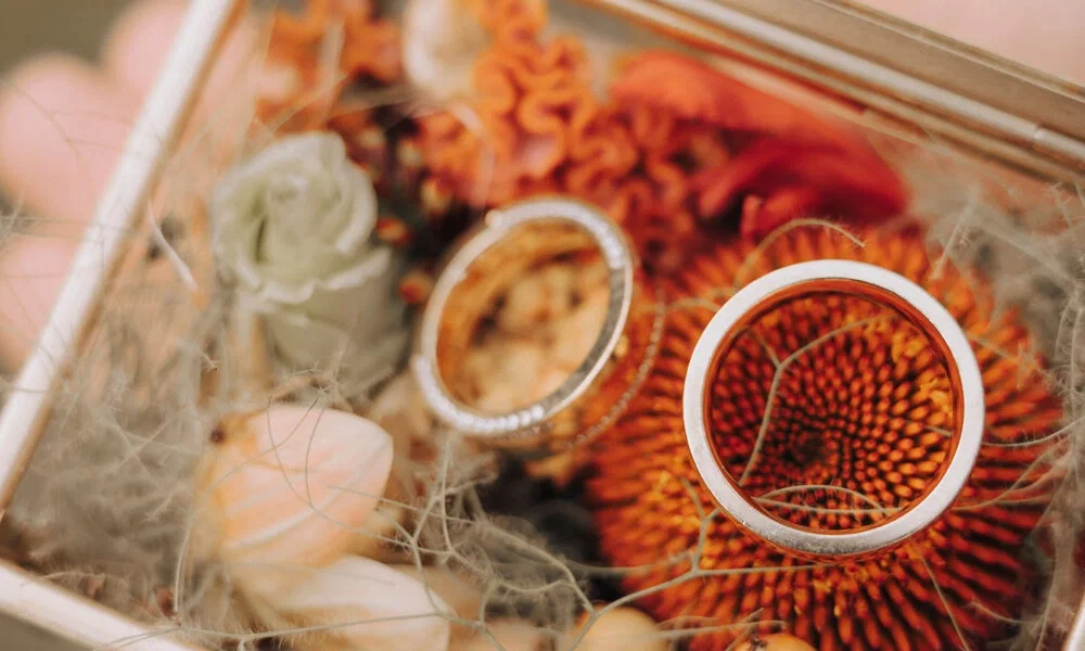 Detailaufnahme von zwei silbernen Trauringen in einem handgefertigten Ringkästchen. Muscheln und orange Blumen sorgen für einen natürlichen und festlichen Rahmen.