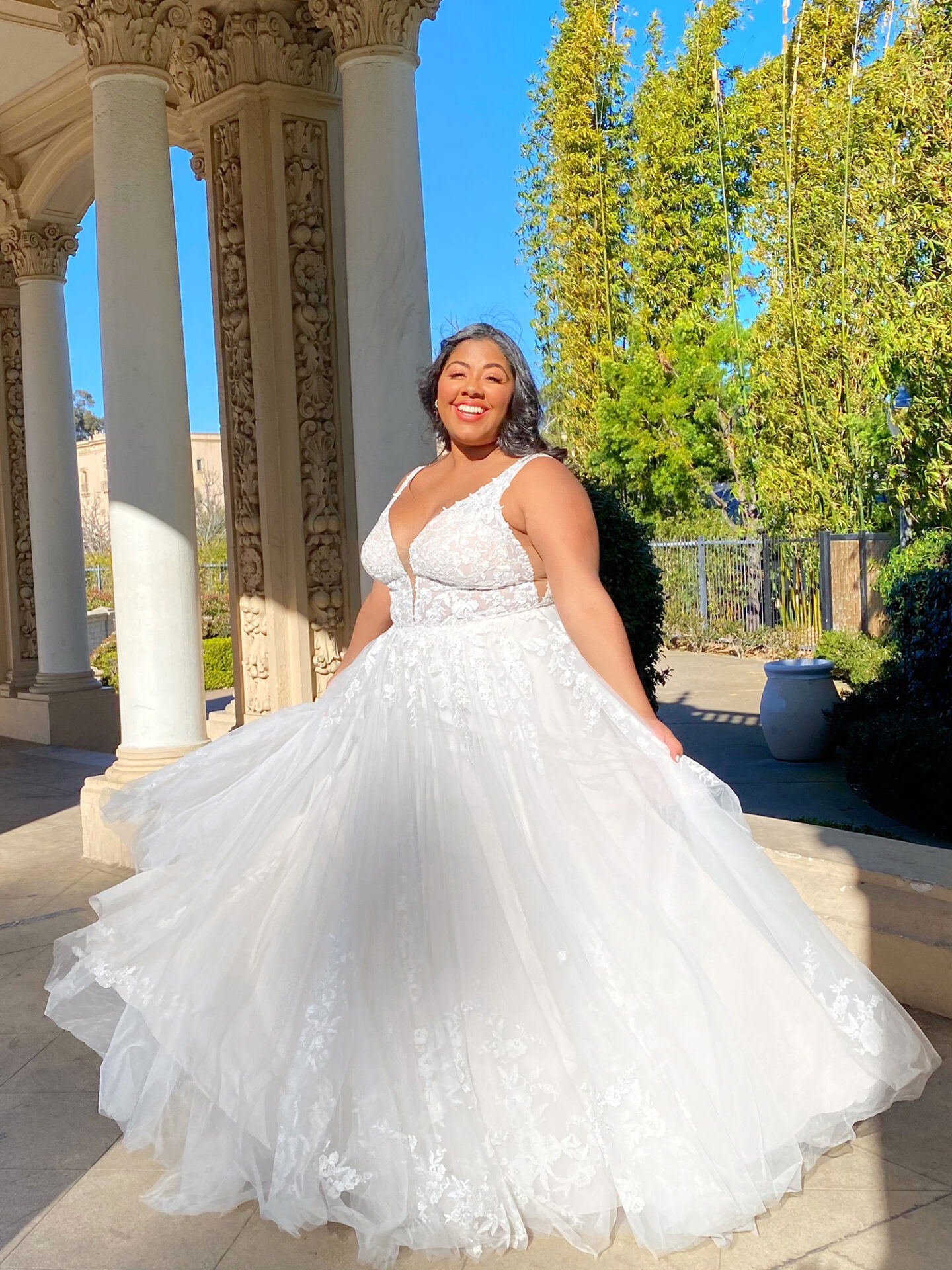 Frau trägt ein florales Brautkleid mit tiefem Ausschnitt und weitem Tüllrock, sie posiert unter antiken Säulen in einer sonnigen Parklandschaft und strahlt Freude aus.