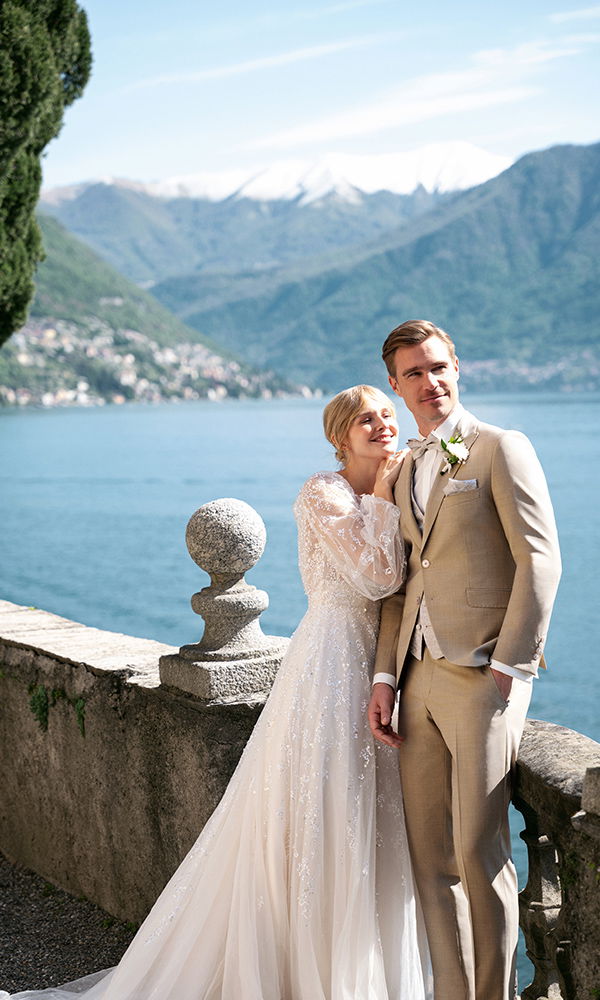 Liebevolle Hochzeitsmomente: Bräutigam in beigem Anzug mit seiner Braut vor Bergpanorama.