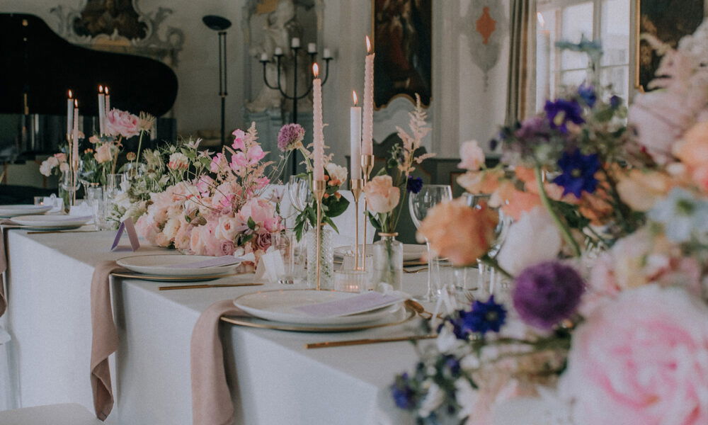 Detailaufnahme einer festlichen Hochzeitstafel, dekoriert mit duftenden Blumen in zarten Rosa- und Lilatönen. Die langen Kerzen und das edle Geschirr runden das stilvolle Ambiente ab.