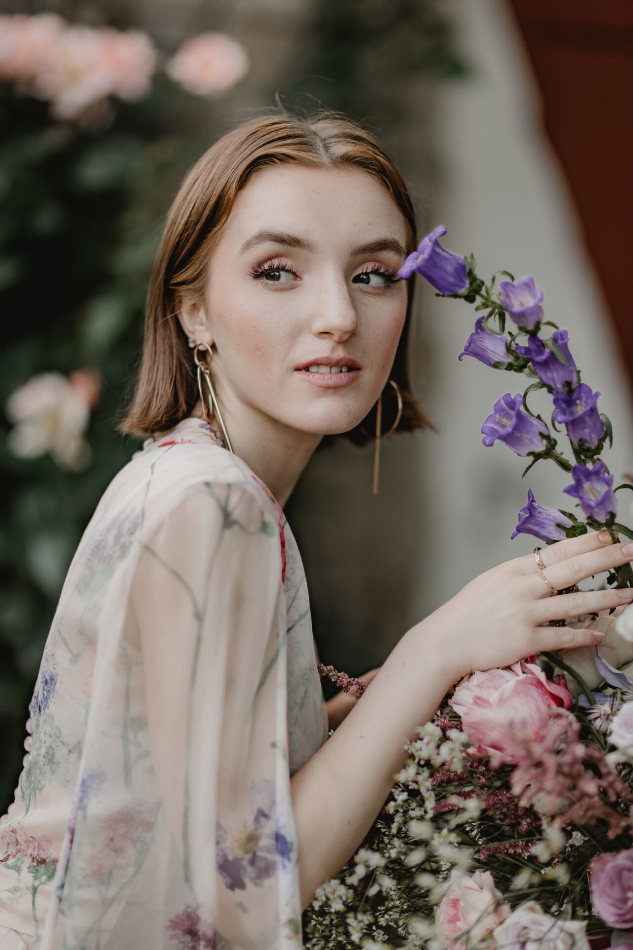 Junge Frau in einem leichten Blumenkleid, die zarte violette Glockenblumen in der Hand hält und seitlich in die Kamera blickt, während Blumen im Hintergrund zu sehen sind.