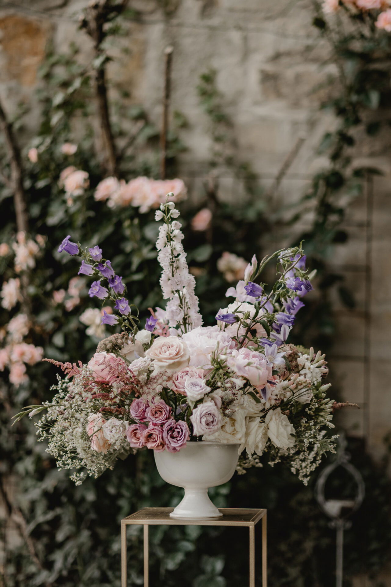 Zierliches Blumenarrangement in einer weißen Vase, bestehend aus verschiedenen Rosensorten und violetten Glockenblumen, dekoriert vor einer bewachsenen Mauer.