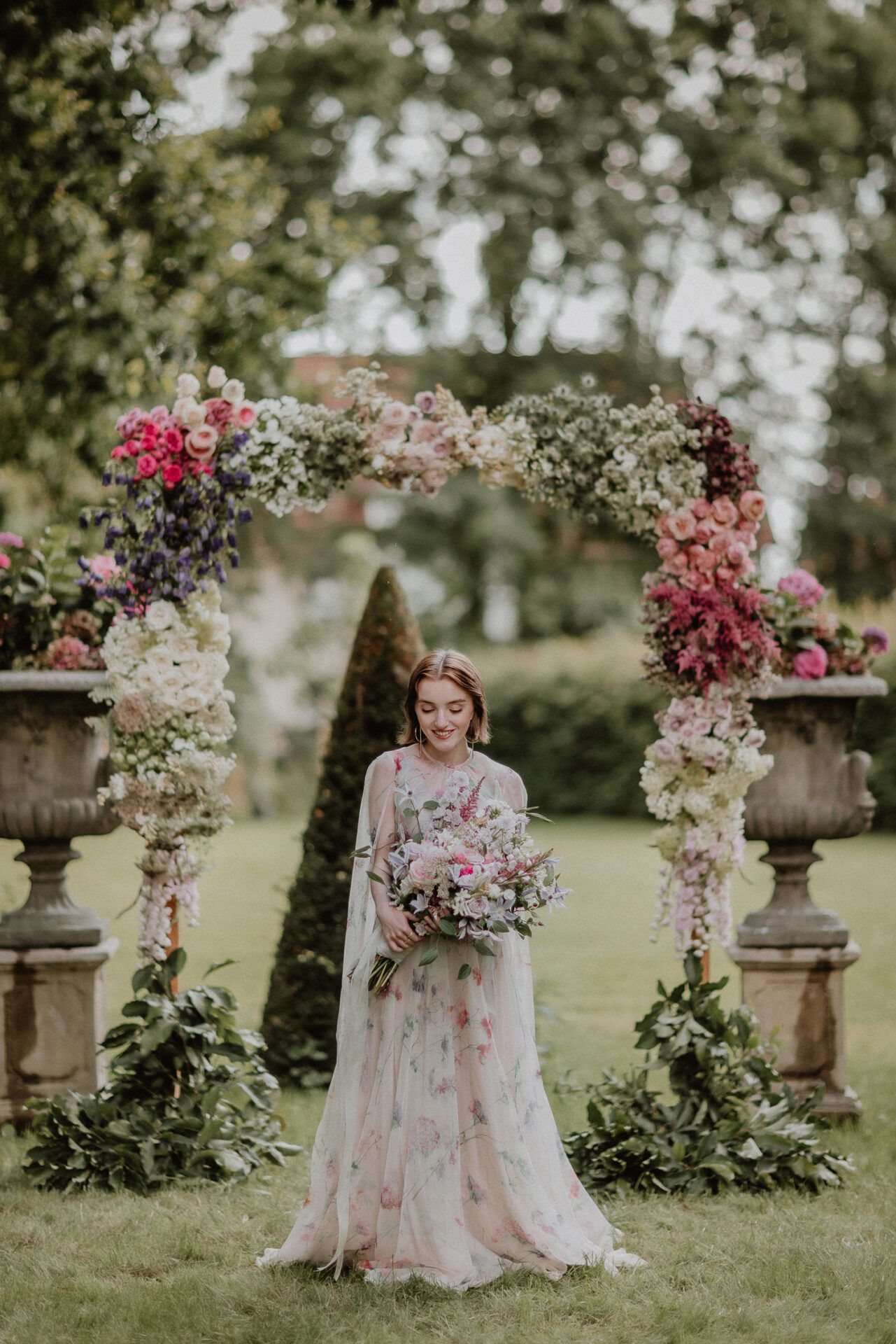 Braut in einem floralen Kleid geht unter einem dekorativen Blumenbogen und hält einen großen Strauß, der perfekt zu der üppigen Naturkulisse des Gartens passt.