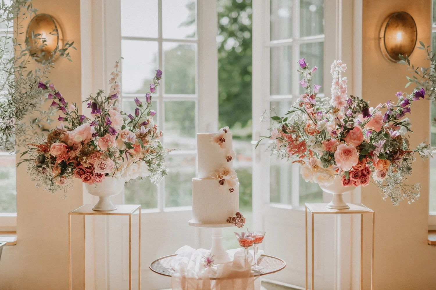 Romantische Hochzeitstischdekoration mit einer schlichten weißen Hochzeitstorte in der Mitte, umgeben von zwei großen Blumenarrangements mit rosa und lila Blüten, vor einer lichtdurchfluteten Fensterfront.