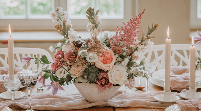 Zierliches Blumengesteck mit Rosen, Astern und Grünpflanzen in zarten Pastelltönen als Mittelpunkt eines dekorativen Hochzeitstisches, der mit Kerzen und elegantem Geschirr gedeckt ist.