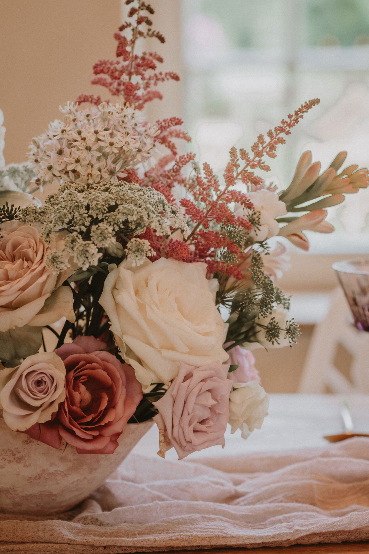 Blumenarrangement in einer Schale mit cremeweißen und rosafarbenen Rosen, umgeben von Wildblumen und pastellfarbenen Akzenten, auf einem Hochzeitstisch mit Leinentuch.