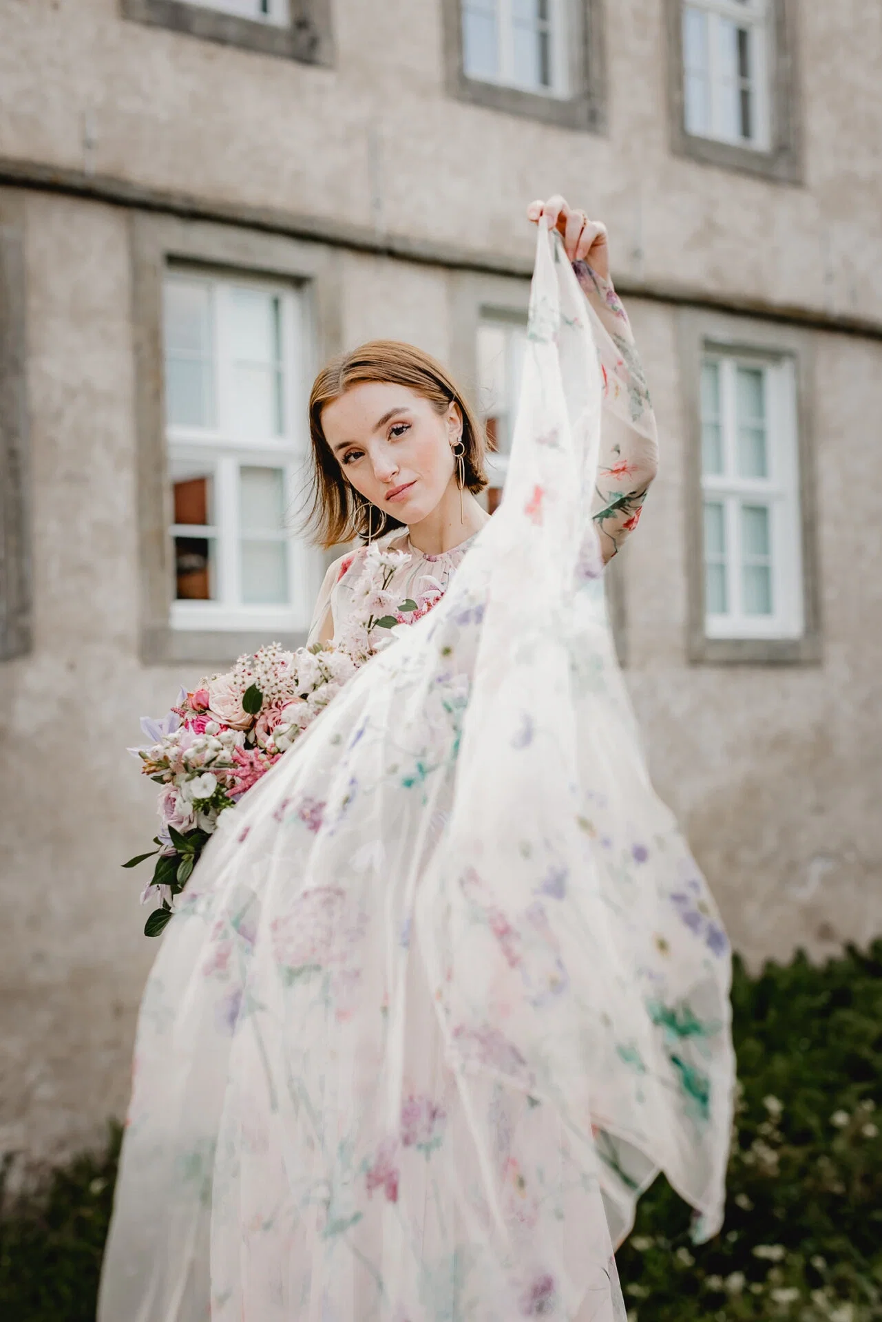 Frau in einem Kleid mit Blumendruck hält das Kleid anmutig zur Seite und posiert mit einem Blumenstrauß vor einem alten Gebäude mit steinernen Wänden.