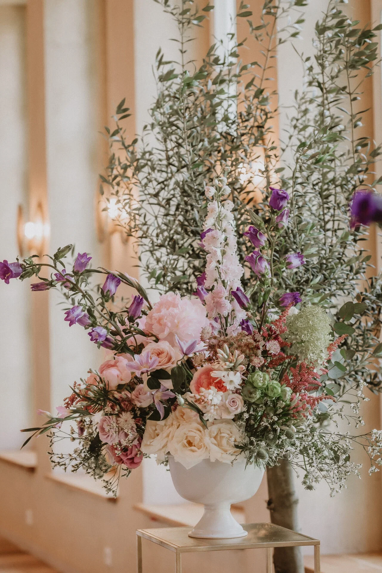 Großes, elegantes Blumenarrangement mit einer Mischung aus lila und rosa Blüten, dekoriert in einer weißen Vase, vor einem eleganten Innenraum mit hohen, verzweigten Pflanzen.