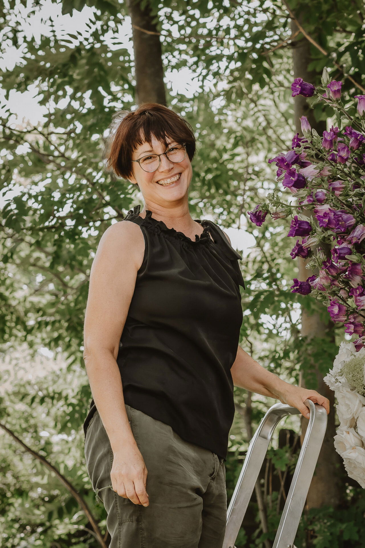 Eine Frau mit einer schwarzen Bluse und Brille posiert entspannt neben einem großen Blumenarrangement mit lila Blüten, während sie im Freien unter Bäumen arbeitet.