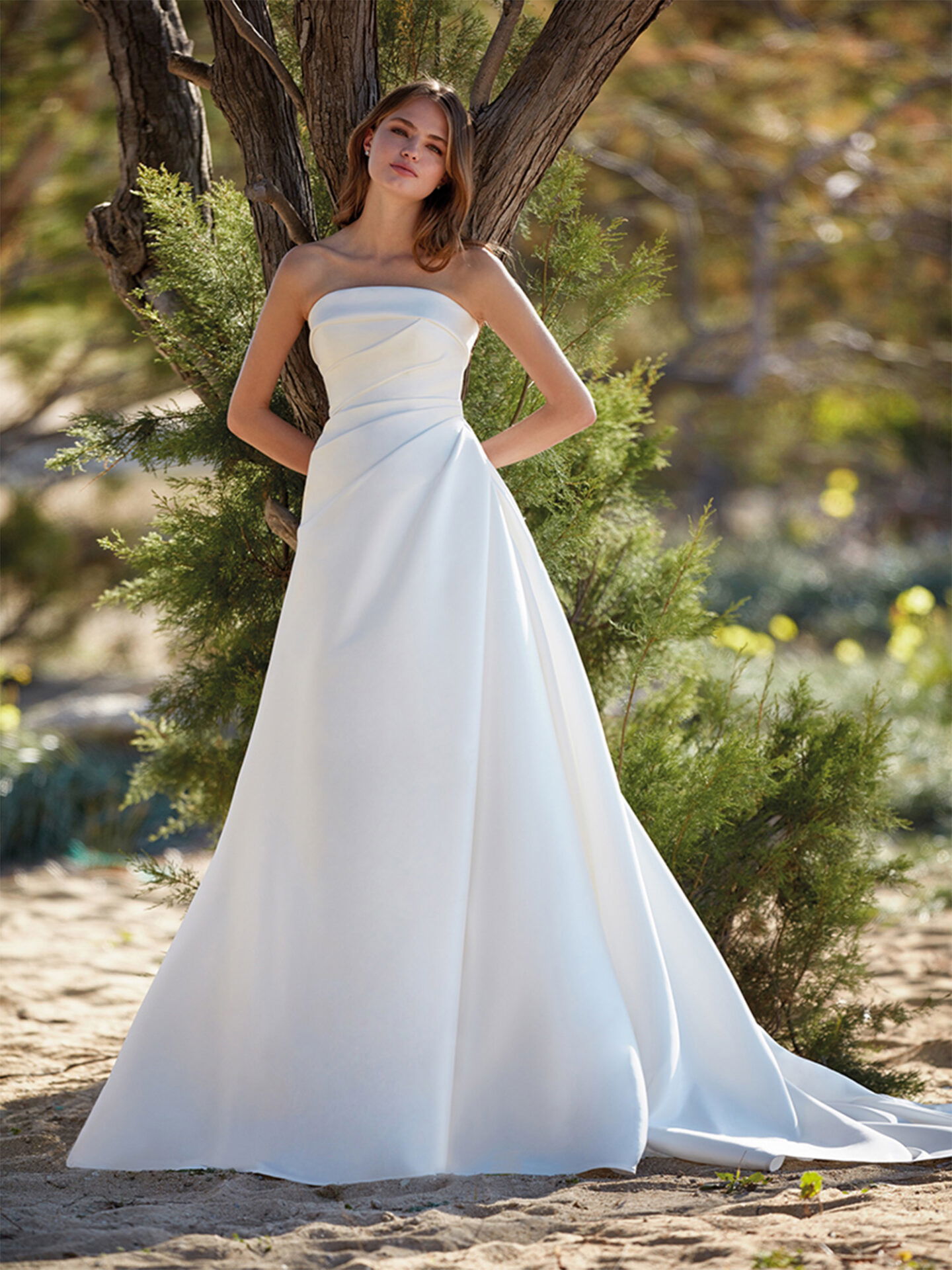 Eine Frau in einem schlichten, eleganten Brautkleid lehnt lässig an einem Baumstamm. Das Kleid mit seiner klaren Linienführung und der fließenden Silhouette passt perfekt zur natürlichen Umgebung aus grünen Büschen und sandigem Boden.