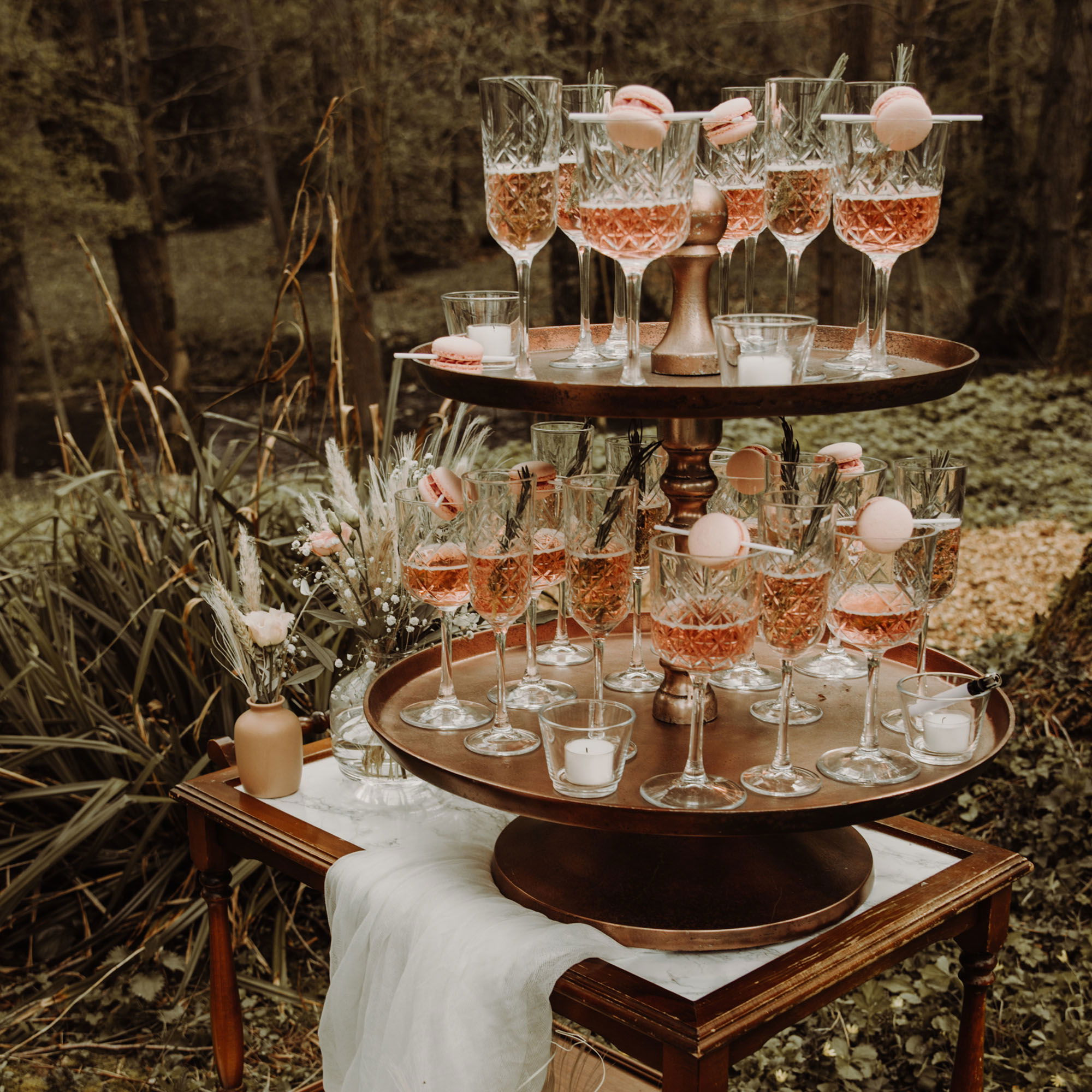 Ein auf zwei Ebenen angeordneter Champagner-Turm, geschmückt mit rosa Macarons, steht auf einem dekorativen Tisch im Freien. Die funkelnden Gläser sind mit Roséwein gefüllt, und das Waldambiente im Hintergrund schafft eine idyllische und luxuriöse Atmosphäre für eine Outdoor-Hochzeitsfeier.