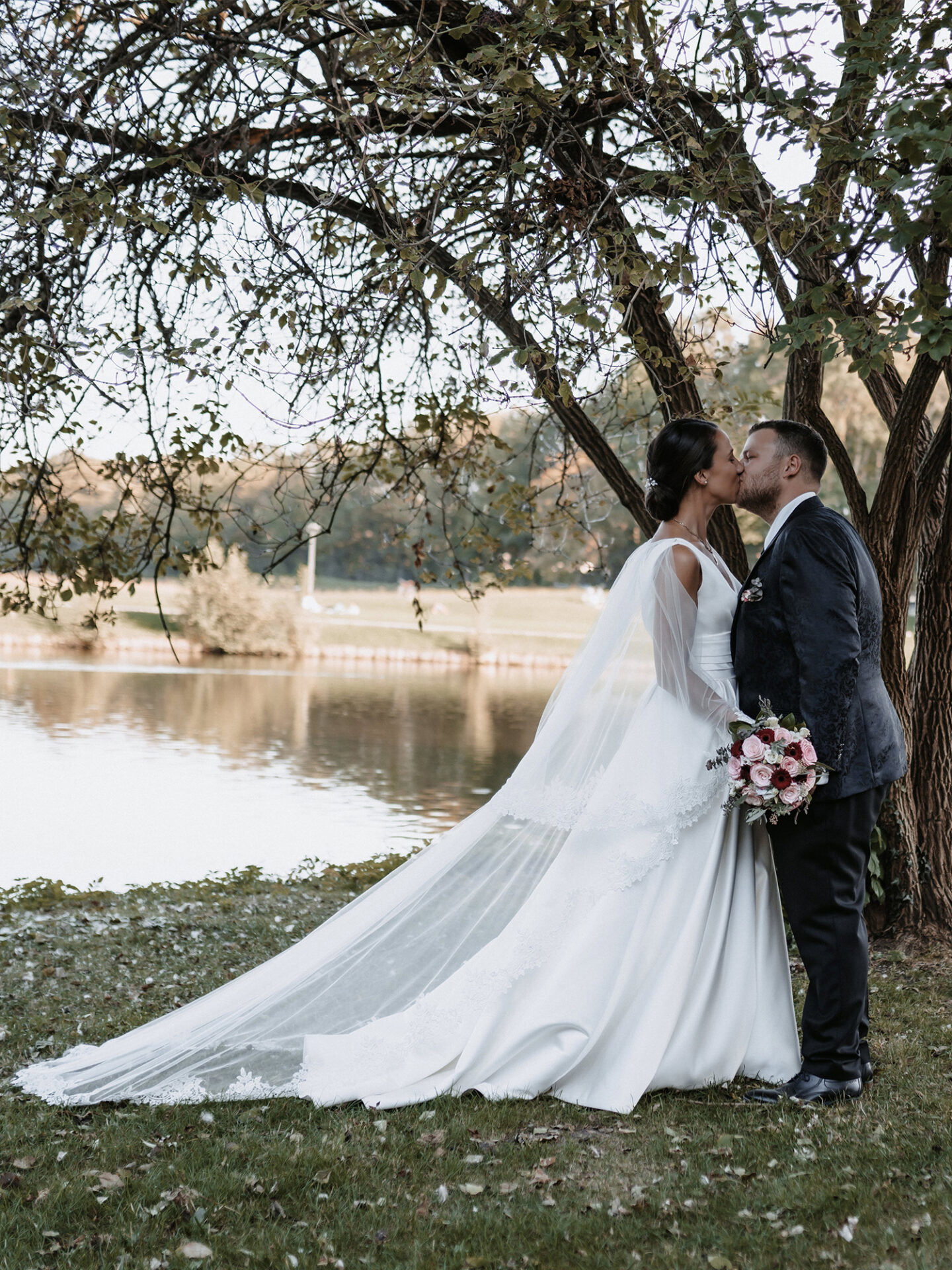 Romantische Szene am Seeufer: Claudia und Hendrik stehen unter einem großen Baum und küssen sich zärtlich. Die Braut trägt ein bodenlanges Kleid mit einem langen, fließenden Schleier, der sich sanft im Wind bewegt. Der Bräutigam ist in einen klassischen dunklen Anzug gekleidet, während die idyllische Natur den Hintergrund bildet.