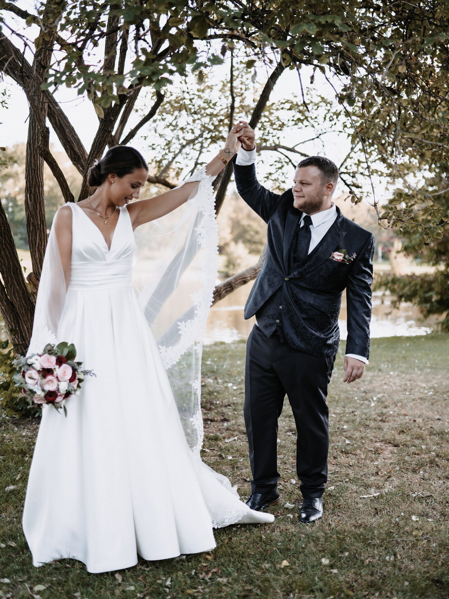 Claudia, in einem klassischen weißen Kleid mit Schleier, hebt lachend ihre Hand, während ihr Ehemann Hendrik sie liebevoll hält. Hendrik trägt einen schwarzen Anzug, und der Hintergrund mit Bäumen und dem See verstärkt die romantische Atmosphäre dieser Hochzeitsaufnahme im Freien.