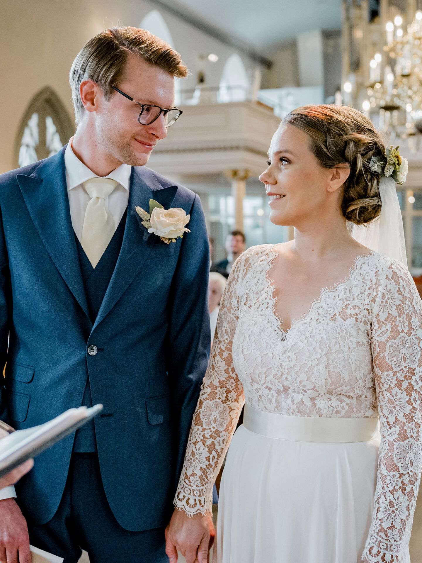 Eine romantische Szene während der Hochzeit, in der sich Braut und Bräutigam tief in die Augen schauen. Die Braut trägt ein Hochzeitskleid mit langen Spitzenärmeln, während der Bräutigam in einem blauen Anzug neben ihr steht. Sie halten sich an den Händen und strahlen Freude aus.