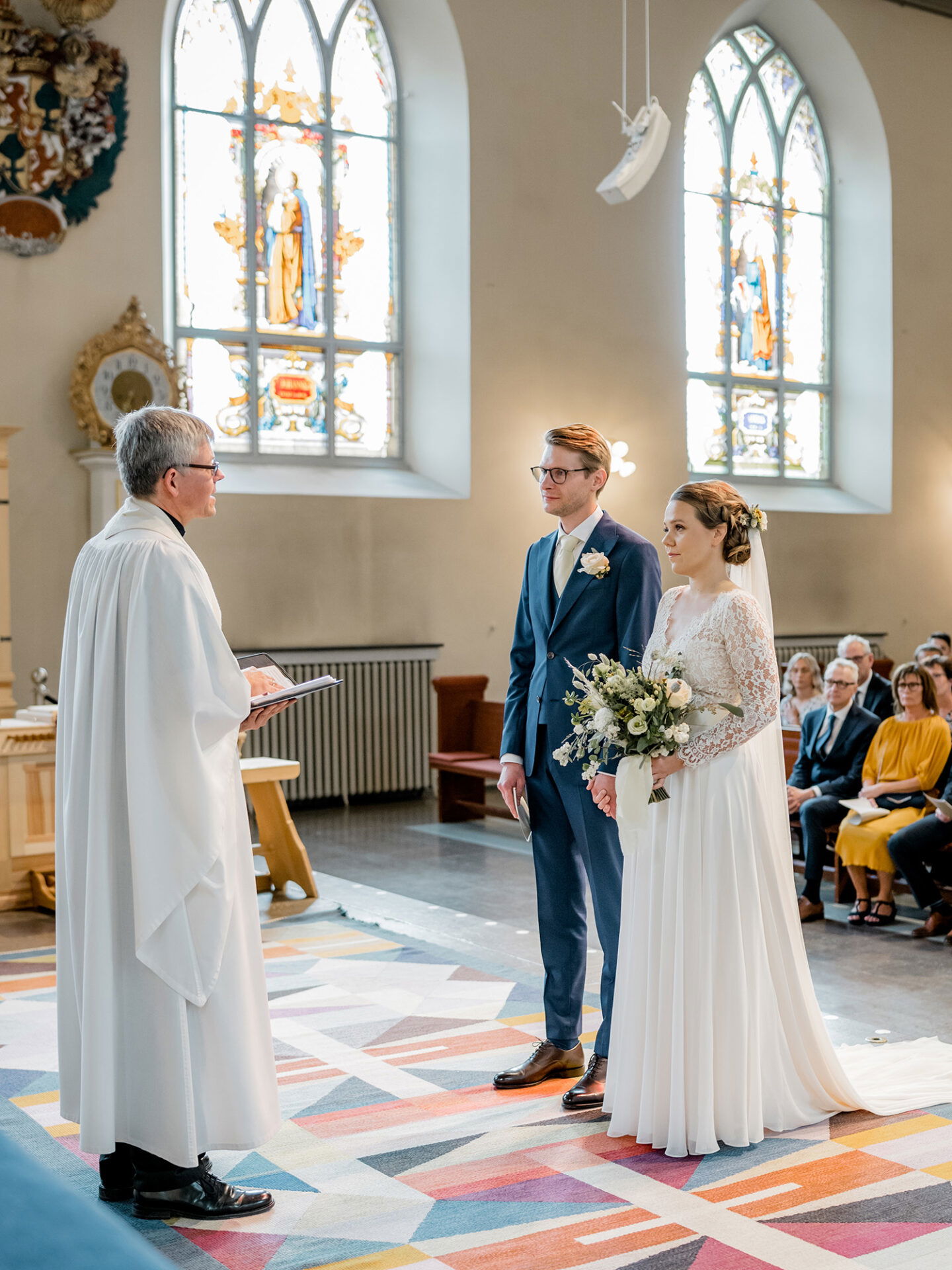 Das Brautpaar, beide in formeller Kleidung, steht in einer Kirche vor einem Priester, der aus einem Buch liest. Die Braut hält ihren Blumenstrauß in der Hand, während sie die Worte des Priesters hören. Die Fenster im Hintergrund sind farbenfroh verziert.