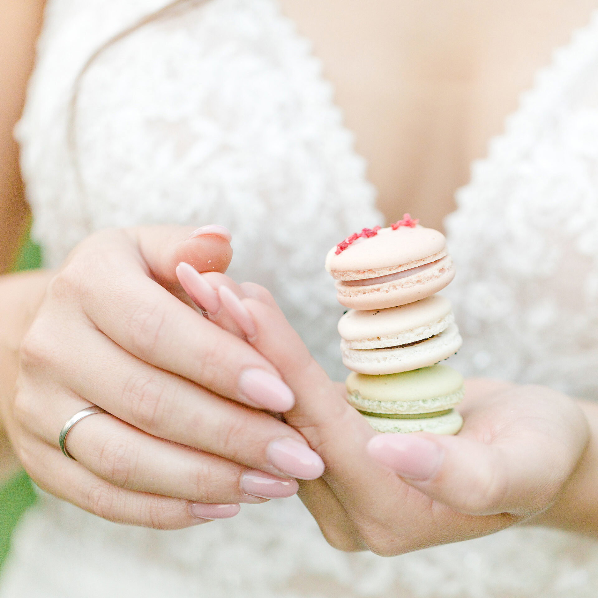 Die Hand einer Braut, die Macarons in verschiedenen Pastellfarben präsentiert, ist in den Fokus gerückt. Ihr fein besticktes Brautkleid verleiht dem Bild eine romantische Atmosphäre.