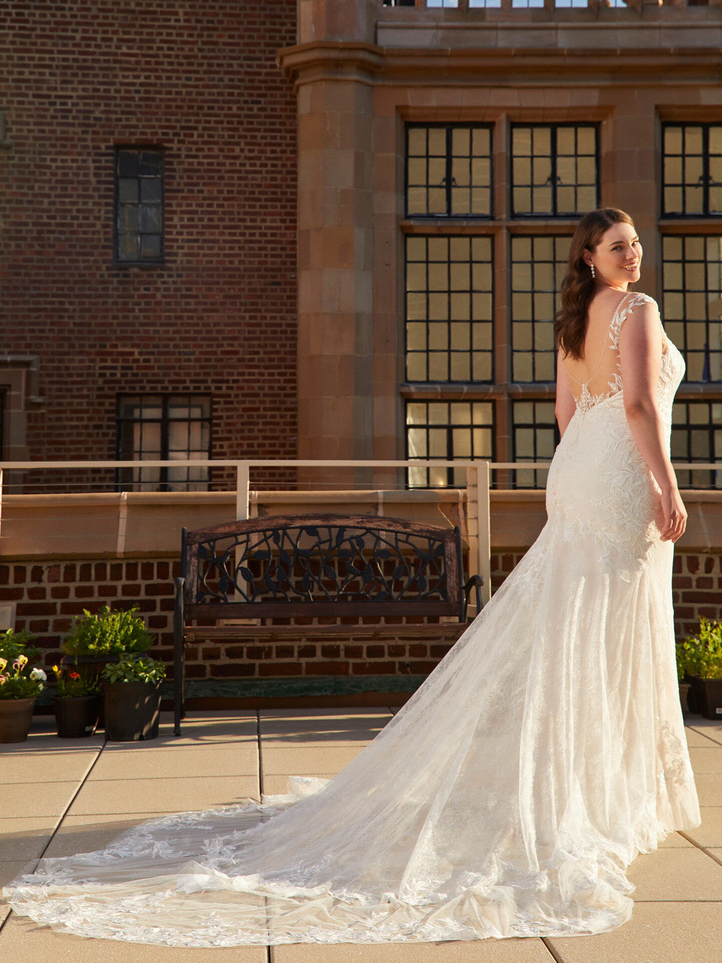 Auf einer sonnigen Terrasse trägt die Braut ein bodenlanges Kleid aus Spitze mit einer beeindruckenden Schleppe, die den Boden sanft berührt. Der Fokus liegt auf dem offenen Rückenausschnitt des Kleides, während die dahinterliegende Backsteinwand und die großen Fenster einen Hauch von urbanem Chic verleihen.