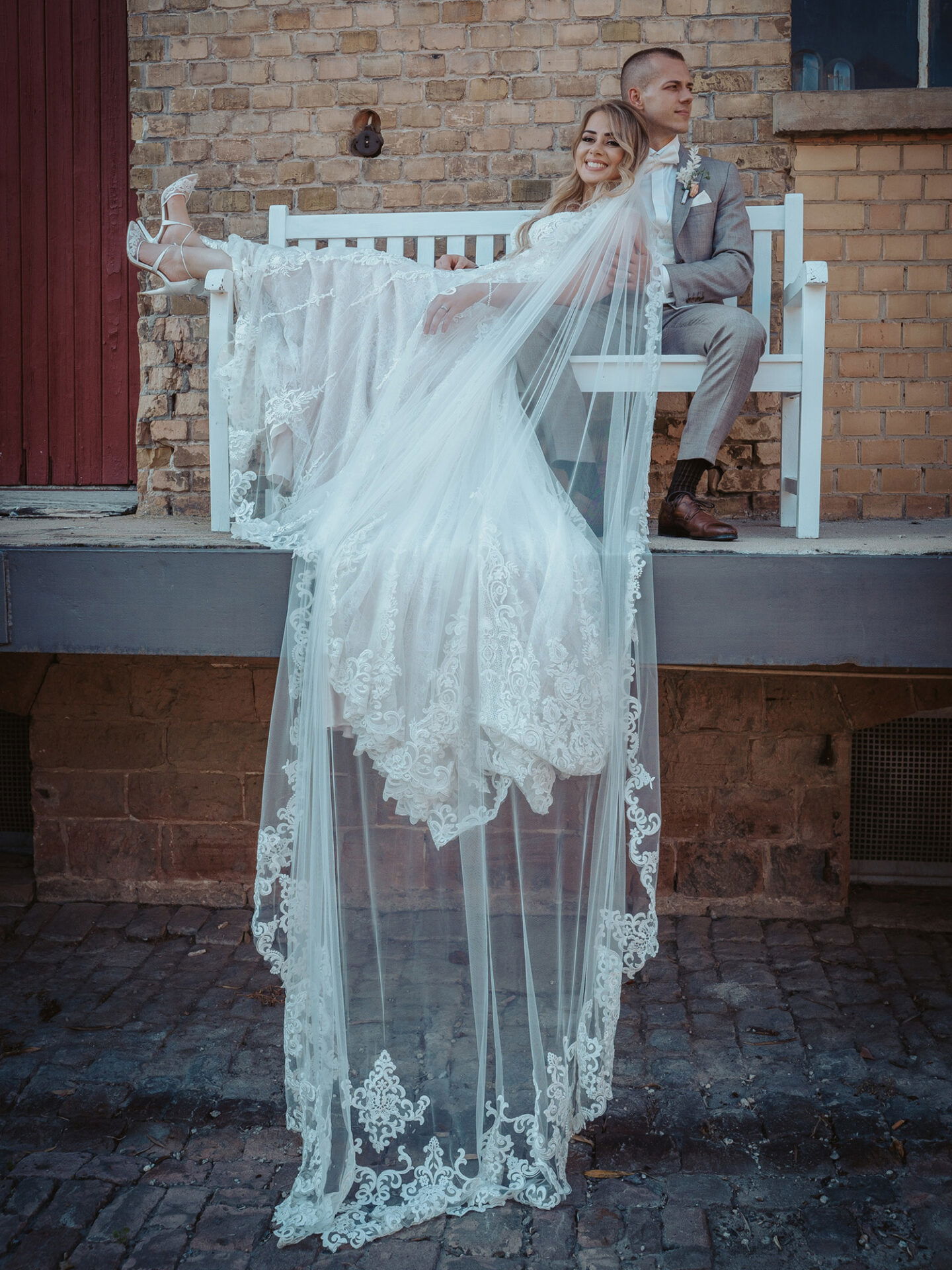 Eine lächelnde Braut liegt entspannt mit ihren Beinen auf einer weißen Bank, ihr langer Schleier fällt über den Rand. Der Bräutigam sitzt hinter ihr und lehnt sich an die Backsteinmauer, während er entspannt zur Seite schaut. Das Paar genießt einen ruhigen Moment nach der Zeremonie.