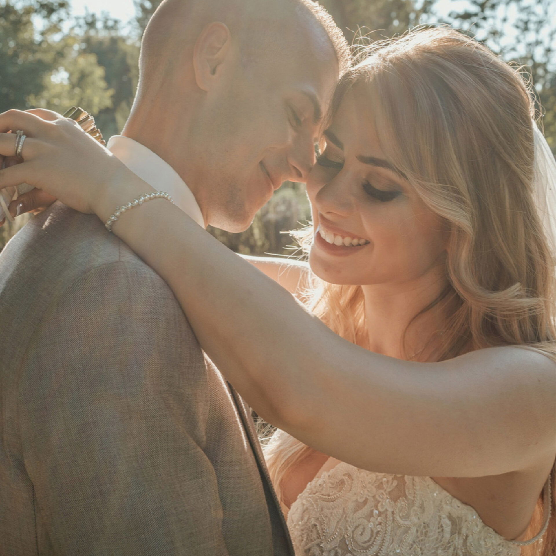 Eine Braut und ein Bräutigam in inniger Umarmung während ihrer Hochzeit. Die Braut trägt ein Detailreiches Brautkleid, und das Paar strahlt Freude aus, während sie im Sonnenlicht eines natürlichen Außenbereichs stehen.