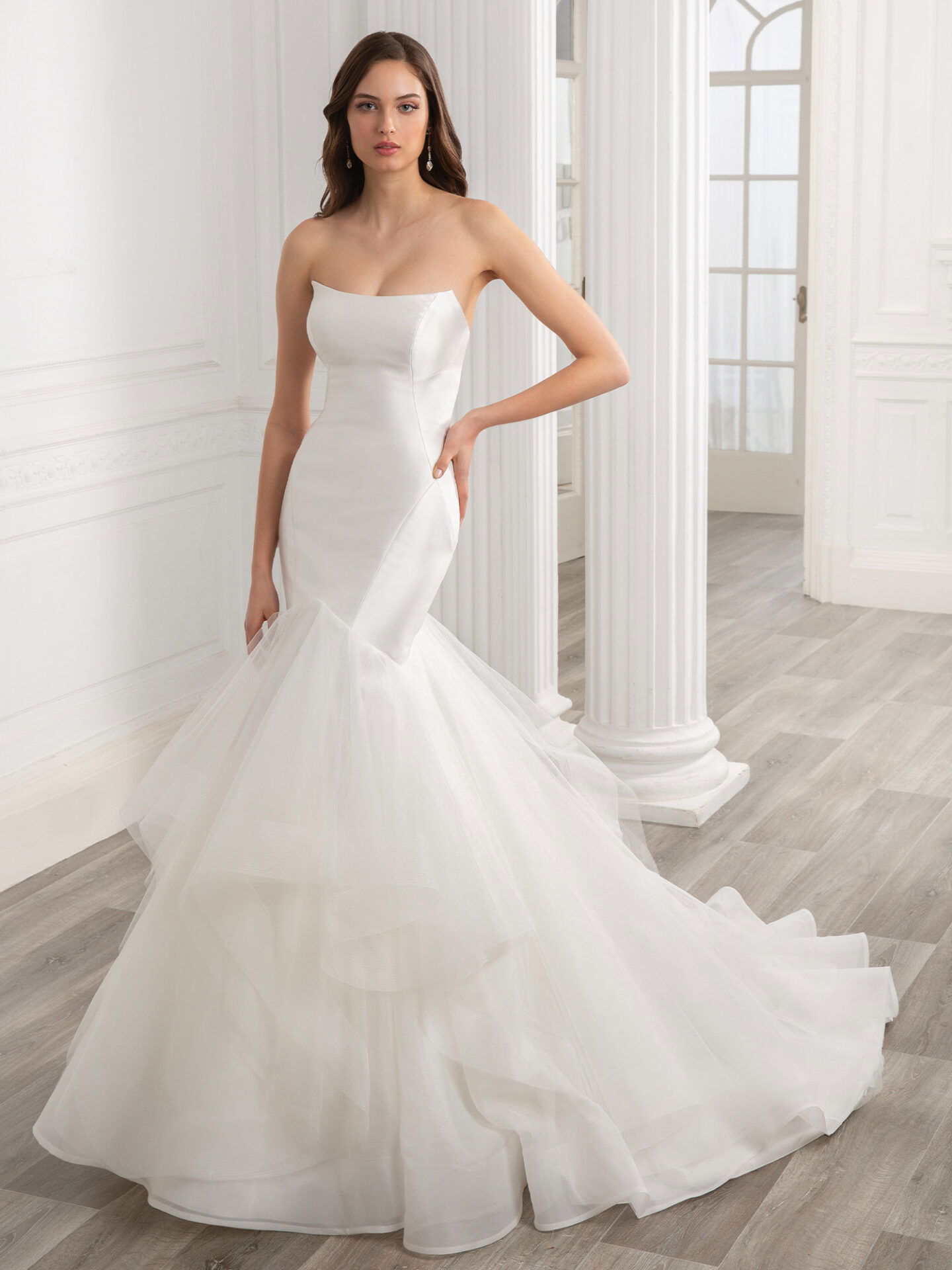 Eine Braut in einem schlichten, aber eleganten, trägerlosen Kleid mit figurbetonter Passform und dramatischem Tüllrock steht vor einer minimalistischen, klassischen Kulisse. Ihr Look ist zeitlos und stilvoll.