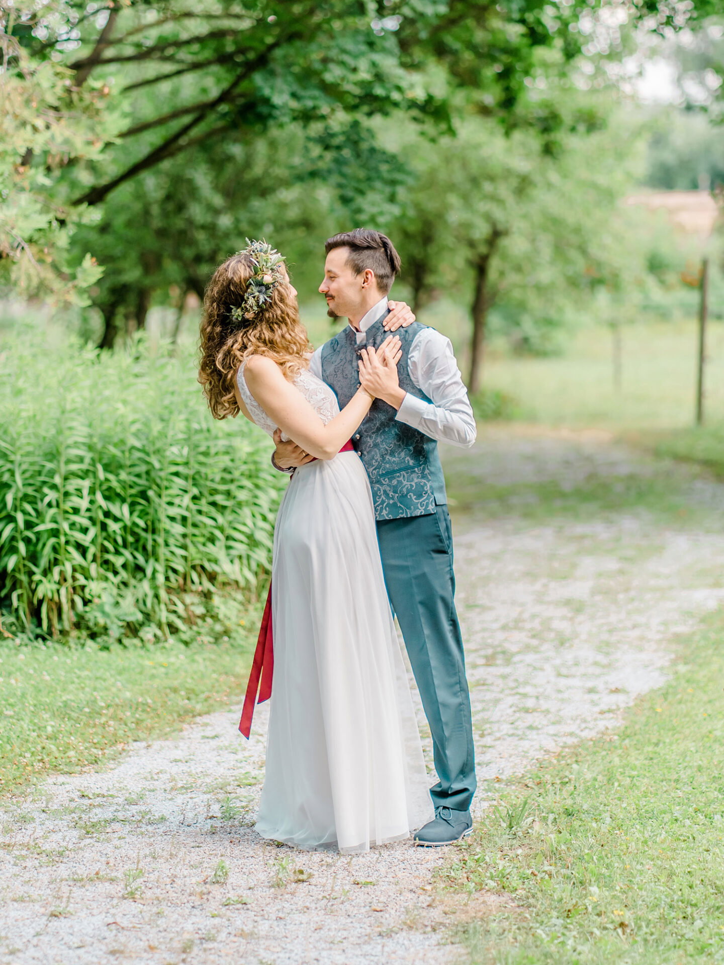 Das Brautpaar genießt einen intimen Moment auf einem naturbelassenen Weg. Die Braut mit einem lockeren Blumenkranz im Haar und der Bräutigam in eleganter Hochzeitsweste scheinen in die Natur vertieft zu sein. Das Paar wirkt inmitten des Grüns und der friedlichen Umgebung völlig glücklich.